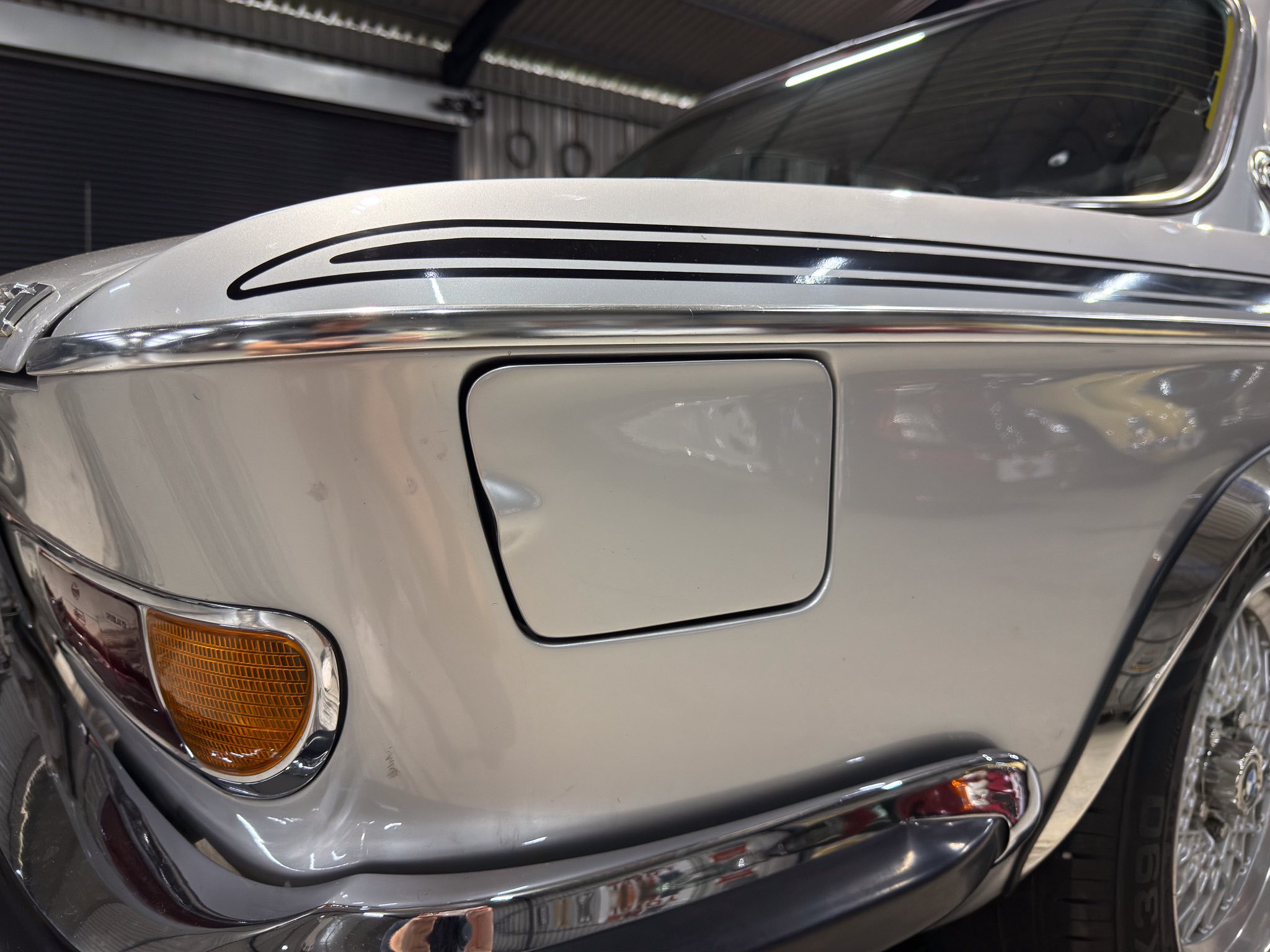 Close-up of a vintage silver car hood with black racing stripes and chrome trim, showing a partial amber front turn signal and windshield edge.