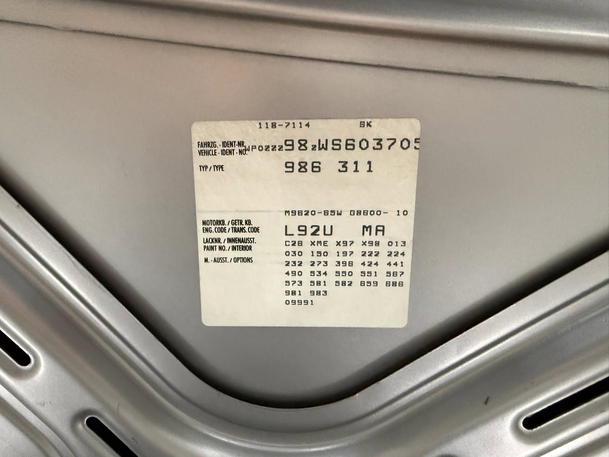 White identification label with multiple alphanumeric codes attached to a grey metal surface ready on a machine part, likely a data/serial plate.