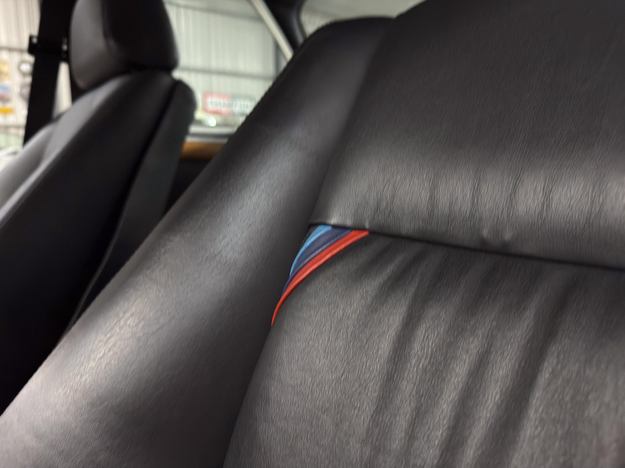 Close-up of a car interior: black leather seat with a blue and red striped seatbelt between the seats.