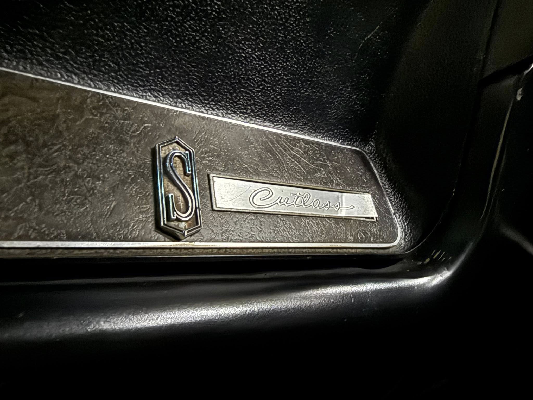 Close-up of a textured black car interior panel with a small hex-edged emblem and a rectangular plate bearing cursive script text.