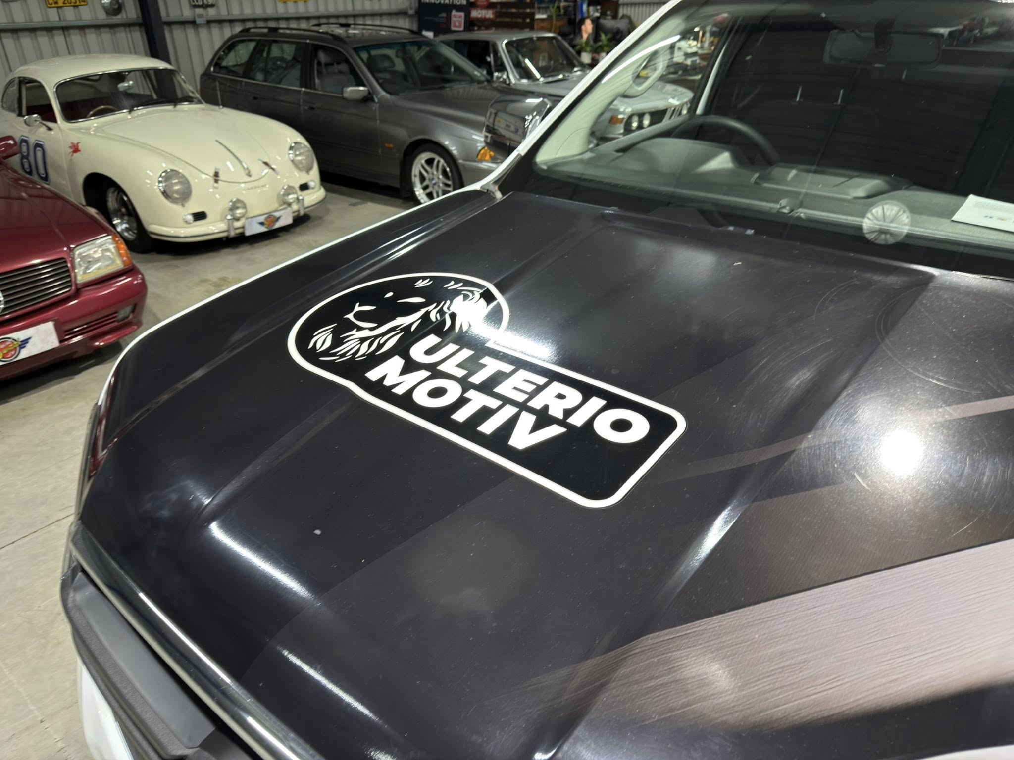 Indoor car showroom with several classic cars, including a white vintage coupe on the left and a dark SUV in the foreground showing a large Ultimate Motor... logo on the hood of the car closest to the camera.