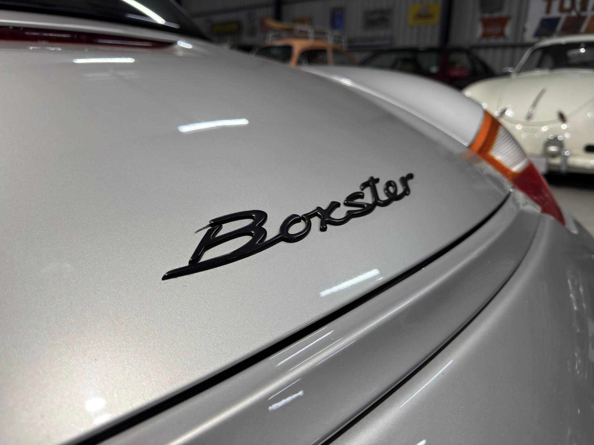 Rear view of a silver Porsche Boxster in a showroom, with other classic cars in the background.