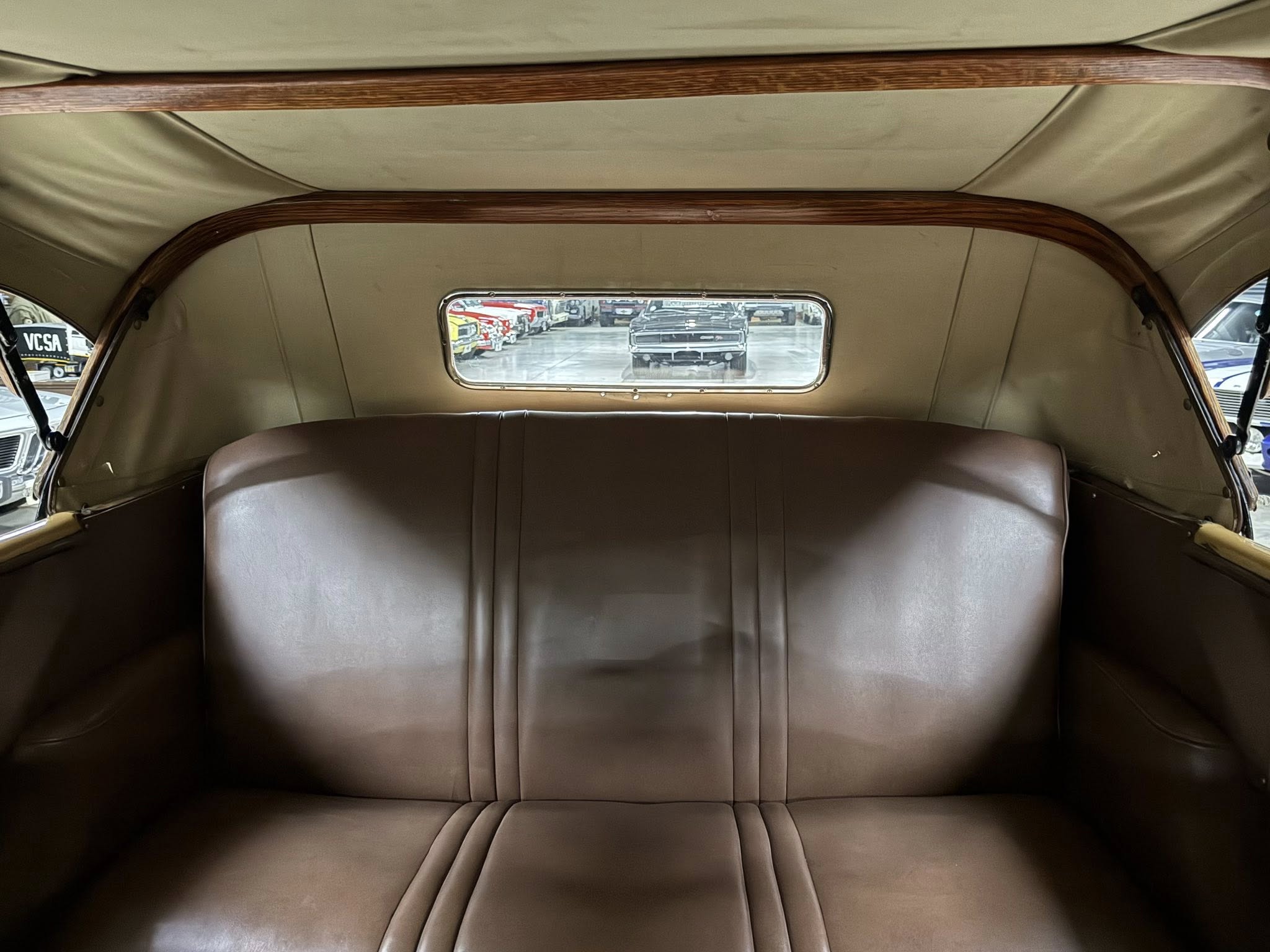 Backseat of a vintage car with beige upholstery and curved wood trim, view of other cars through the rear window.