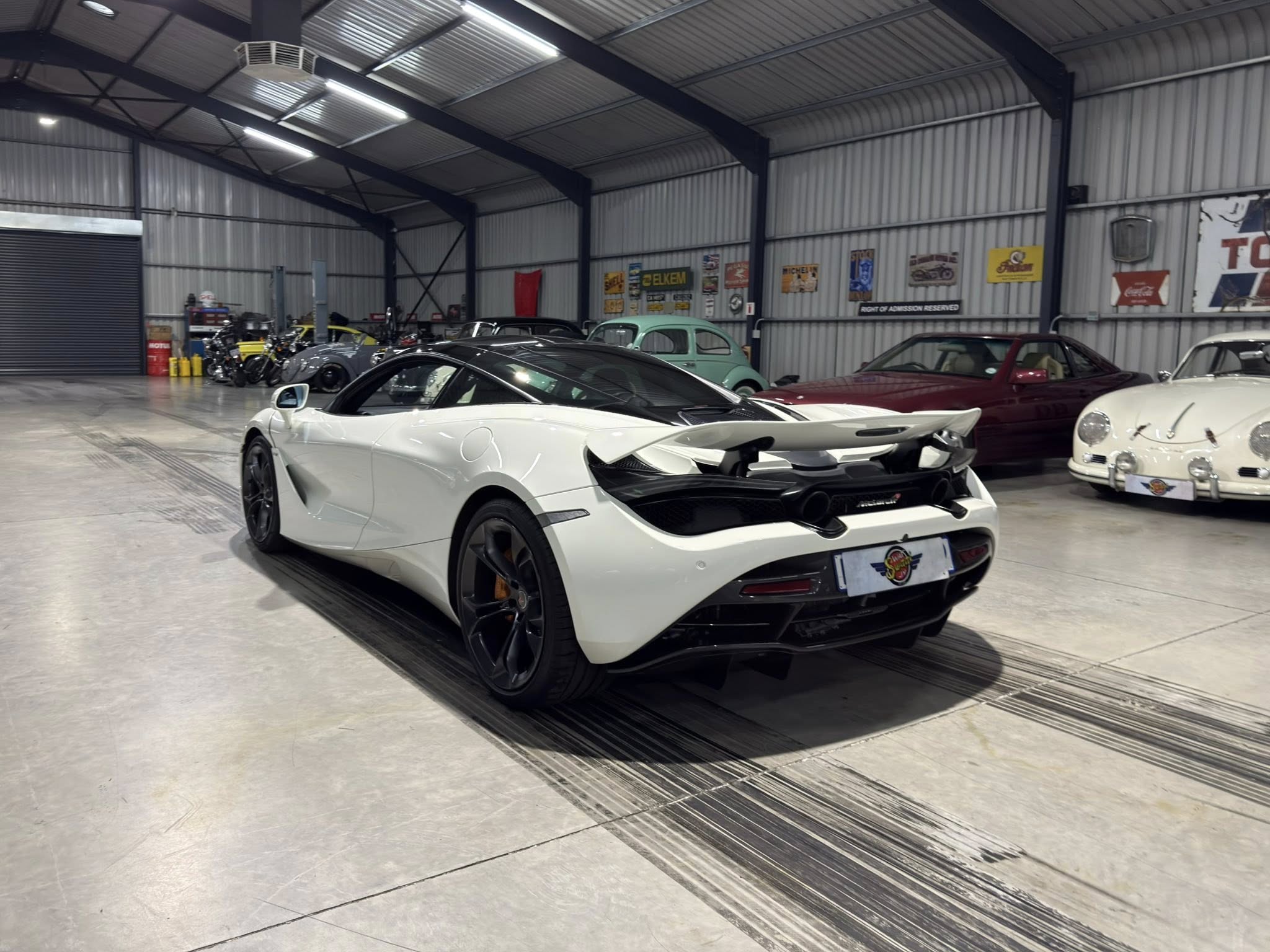 Inside a well-lit indoor showroom with a white sports car in the foreground and vintage cars along the back wall.