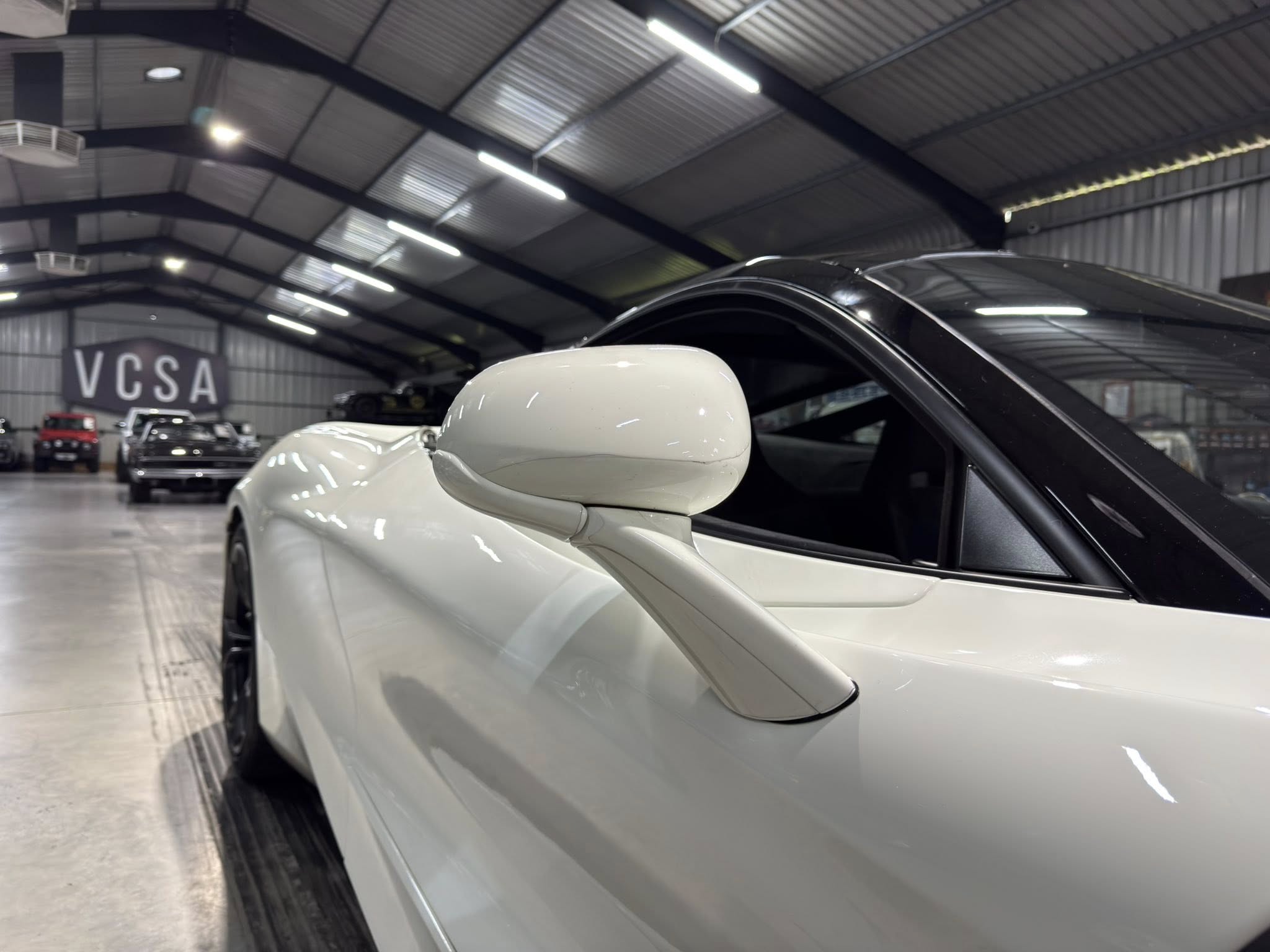 Close-up of a white car's side mirror and door pane inside a showroom, with other vehicles and a large VCSA sign in the background.