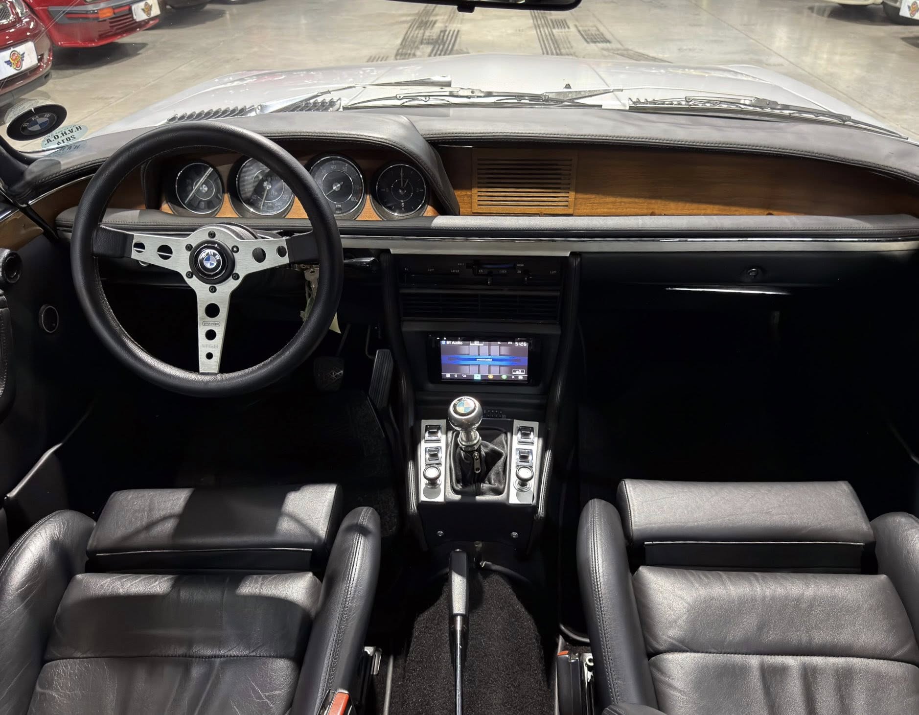 Interior of a classic BMW with black leather seats, wood dashboard trim, and a three‑spoke metal steering wheel.