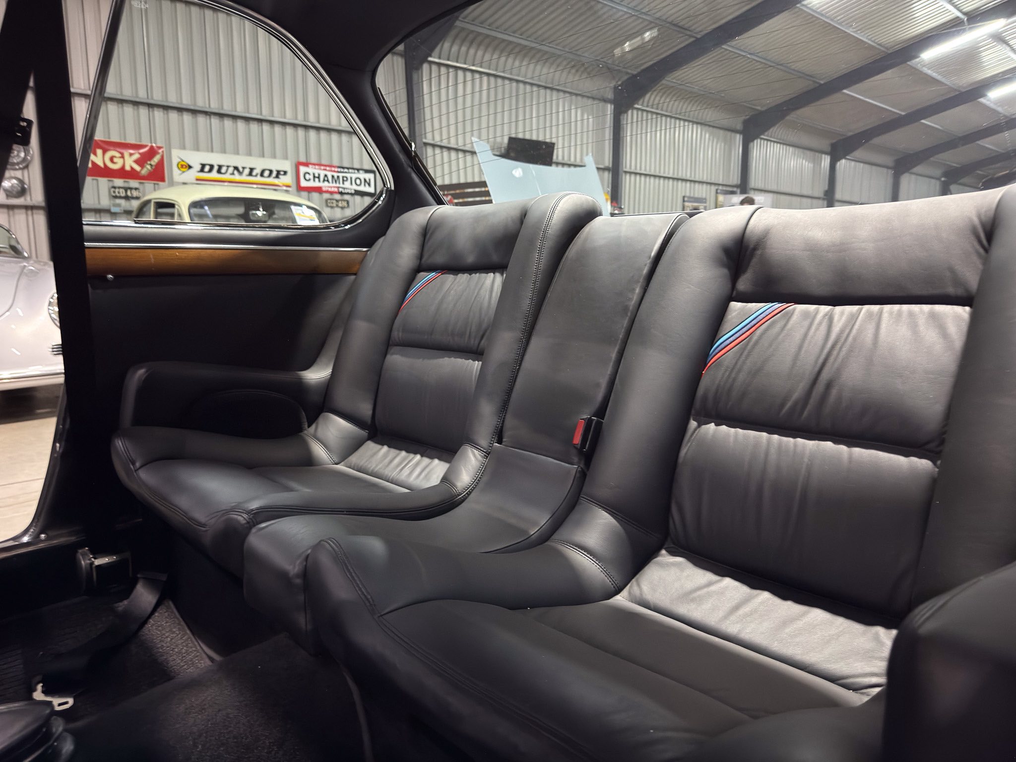 Rear passenger seats in a black leather car interior, inside a showroom with a white vintage car and banners visible through the window.