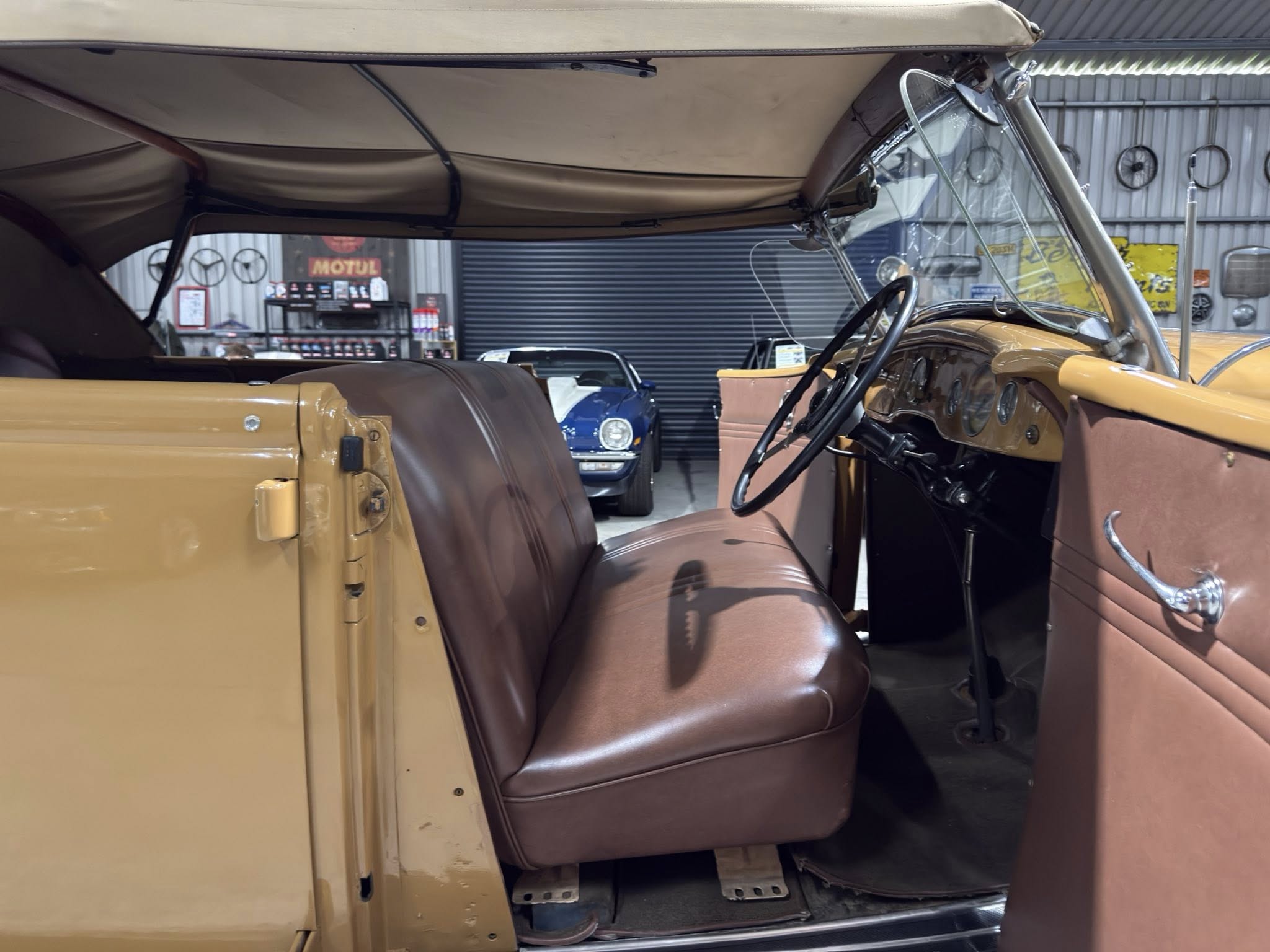 Inside vintage car showing tan leather benches and steering wheel, in a workshop with other vehicles in the background.