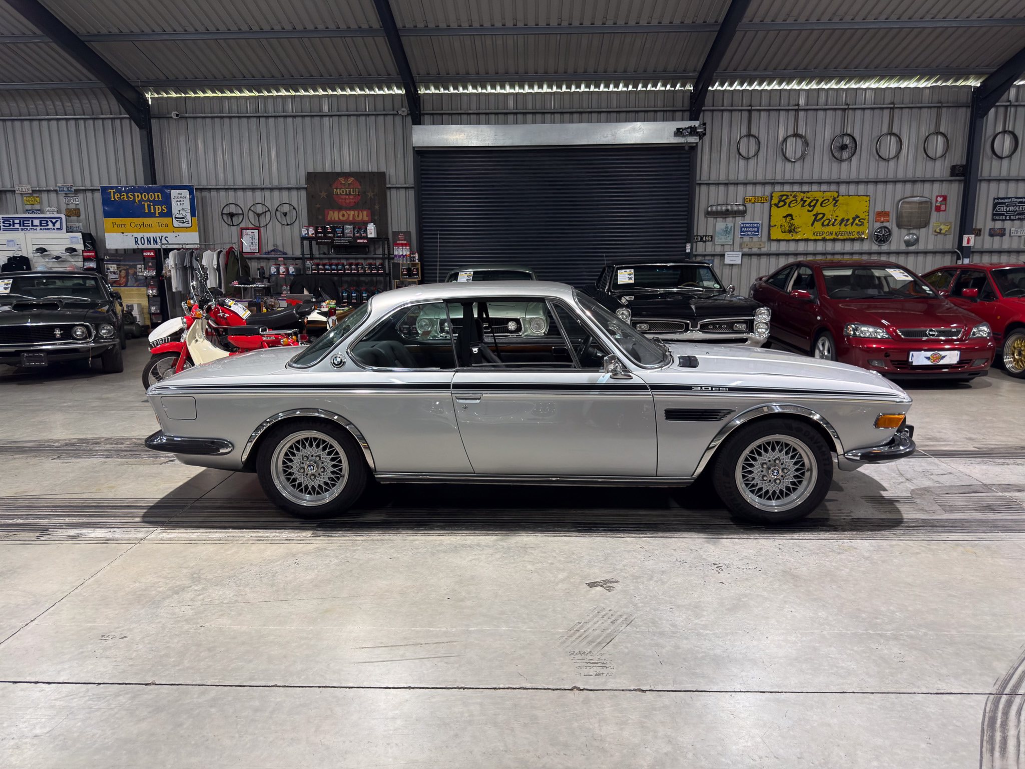 Inside a retro car showroom, a silver vintage coupe is parked in the foreground with several classic cars lined up behind it and vintage signs on the metal wall.