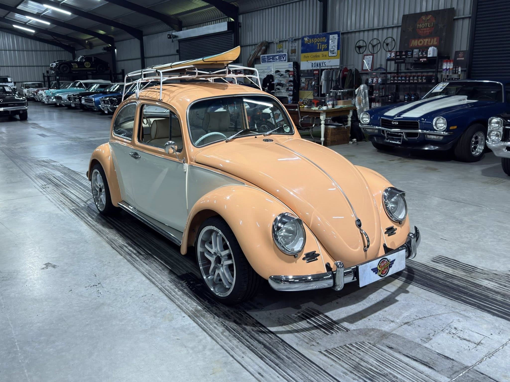 Front view of a peach-colored vintage VW Beetle with a roof rack and surfboard, inside a classic car showroom.