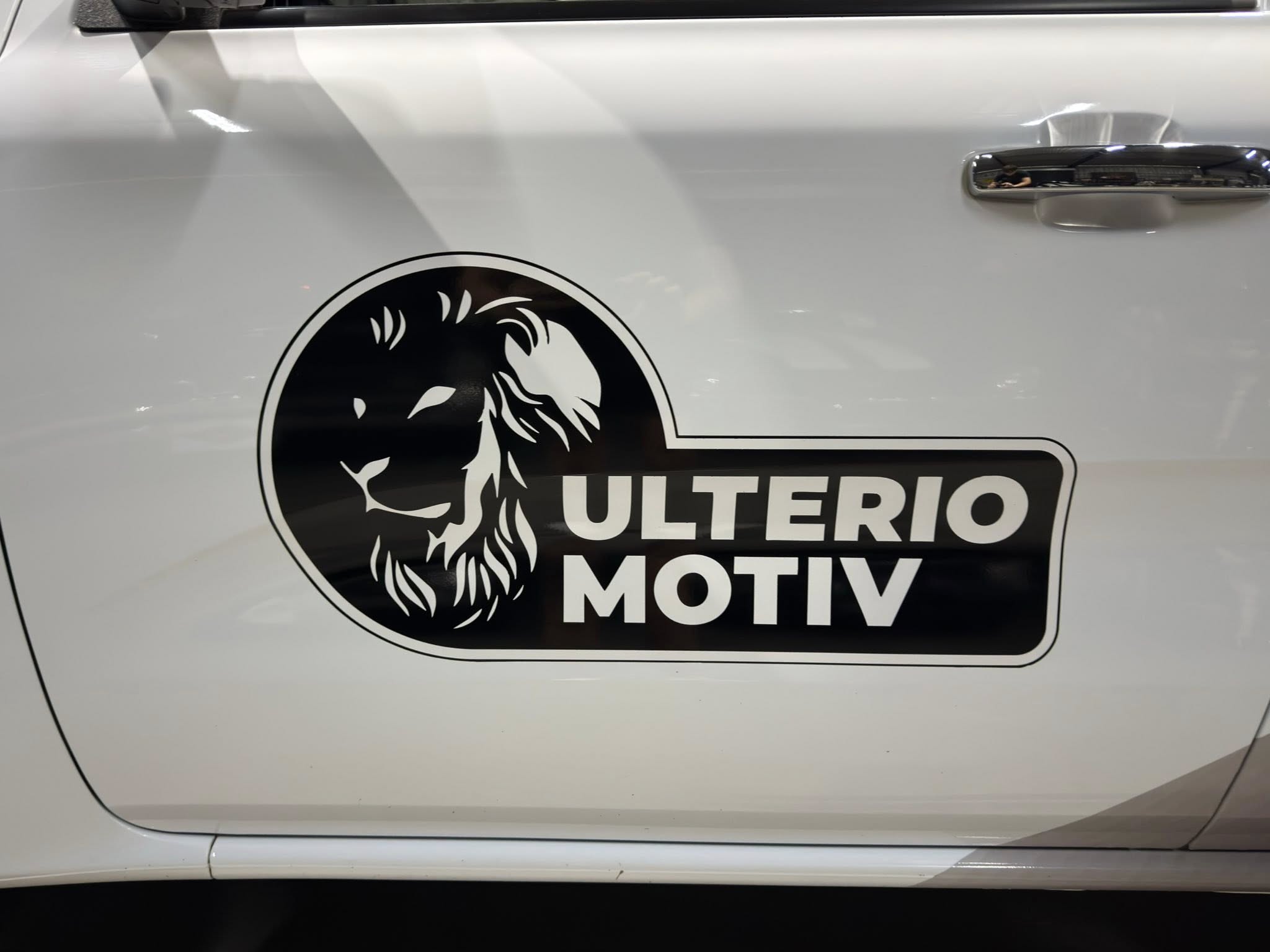 Close-up of a white vehicle door with a circular black logo featuring a stylized lion and the words 'ULTERIO MOTIV' (partially visible).