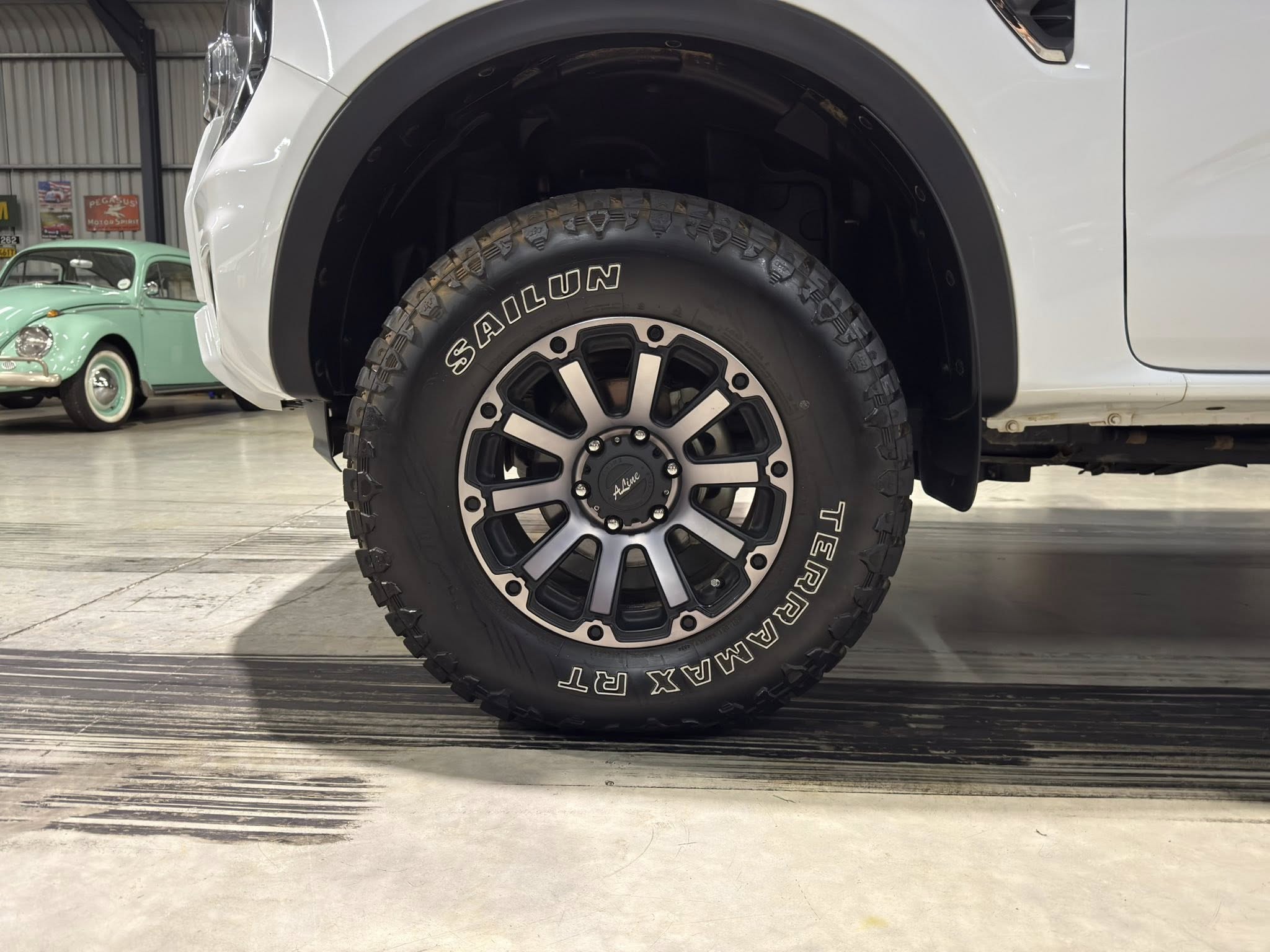 Close-up of a white pickup's large off-road tire and alloy wheel, indoors with a mint-green vintage car in the background.