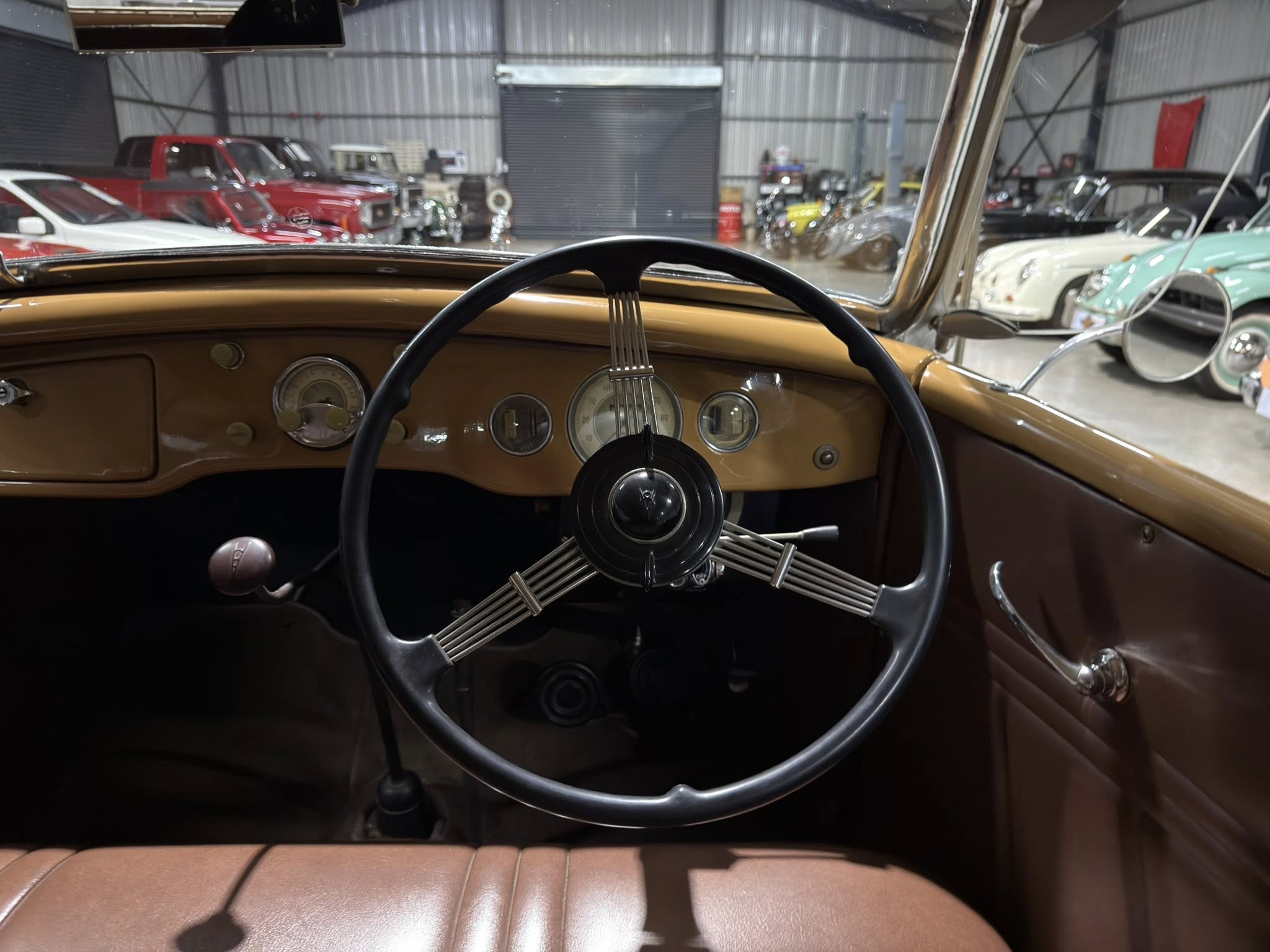 View from the driver's seat of a brown vintage car, showing the steering wheel and classic gauges with a car showroom in the background.