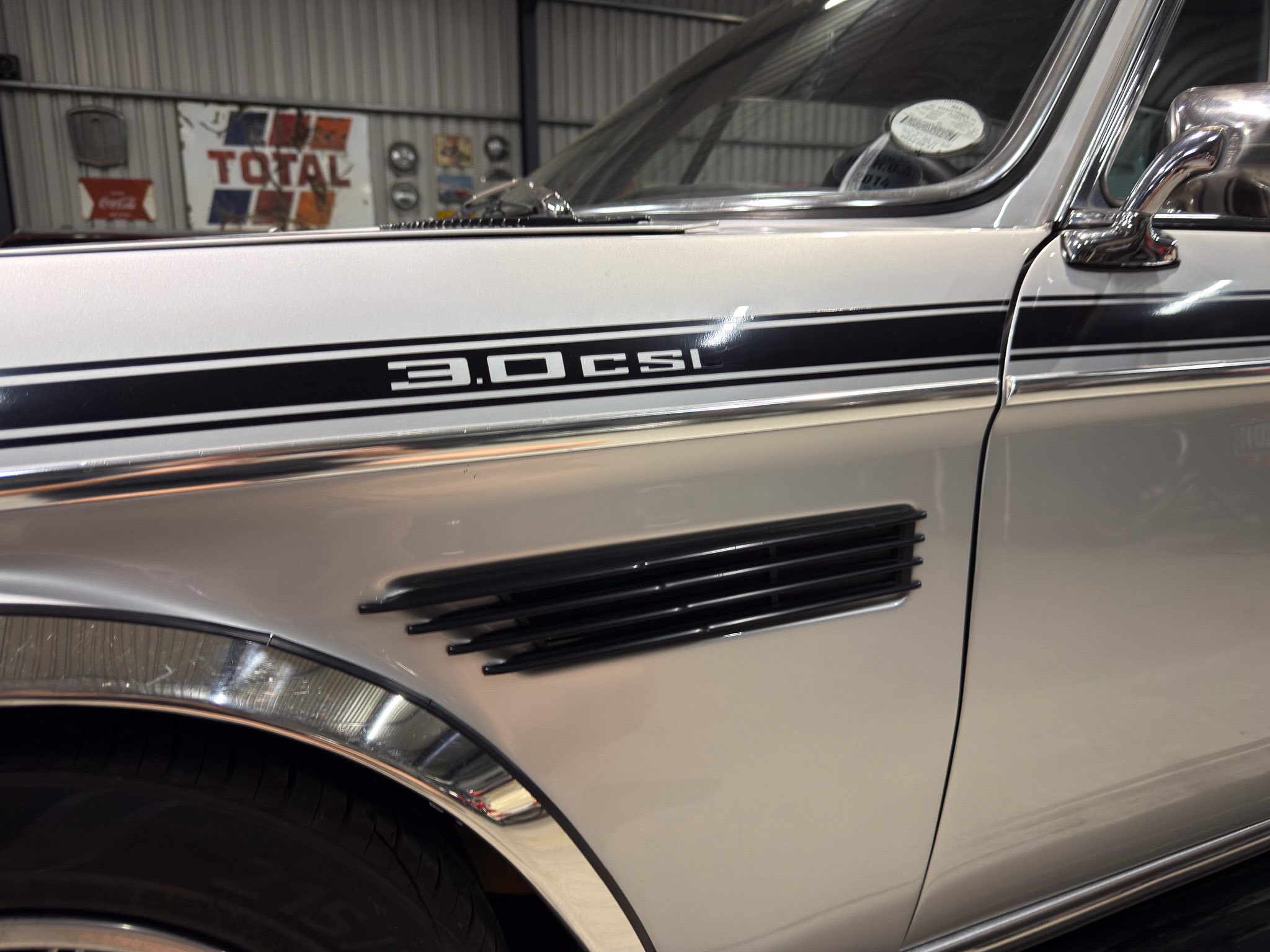 Close-up of a classic silver car side panel with chrome trim and the badge reading '3.0 CSI' or '3.0CSI' along the lower body line.