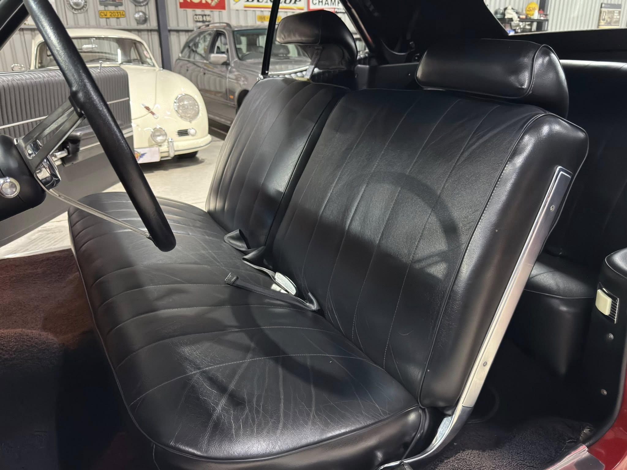Black leather car seats and steering wheel inside a classic car, with vintage cars visible in the background in a showroom or garage.