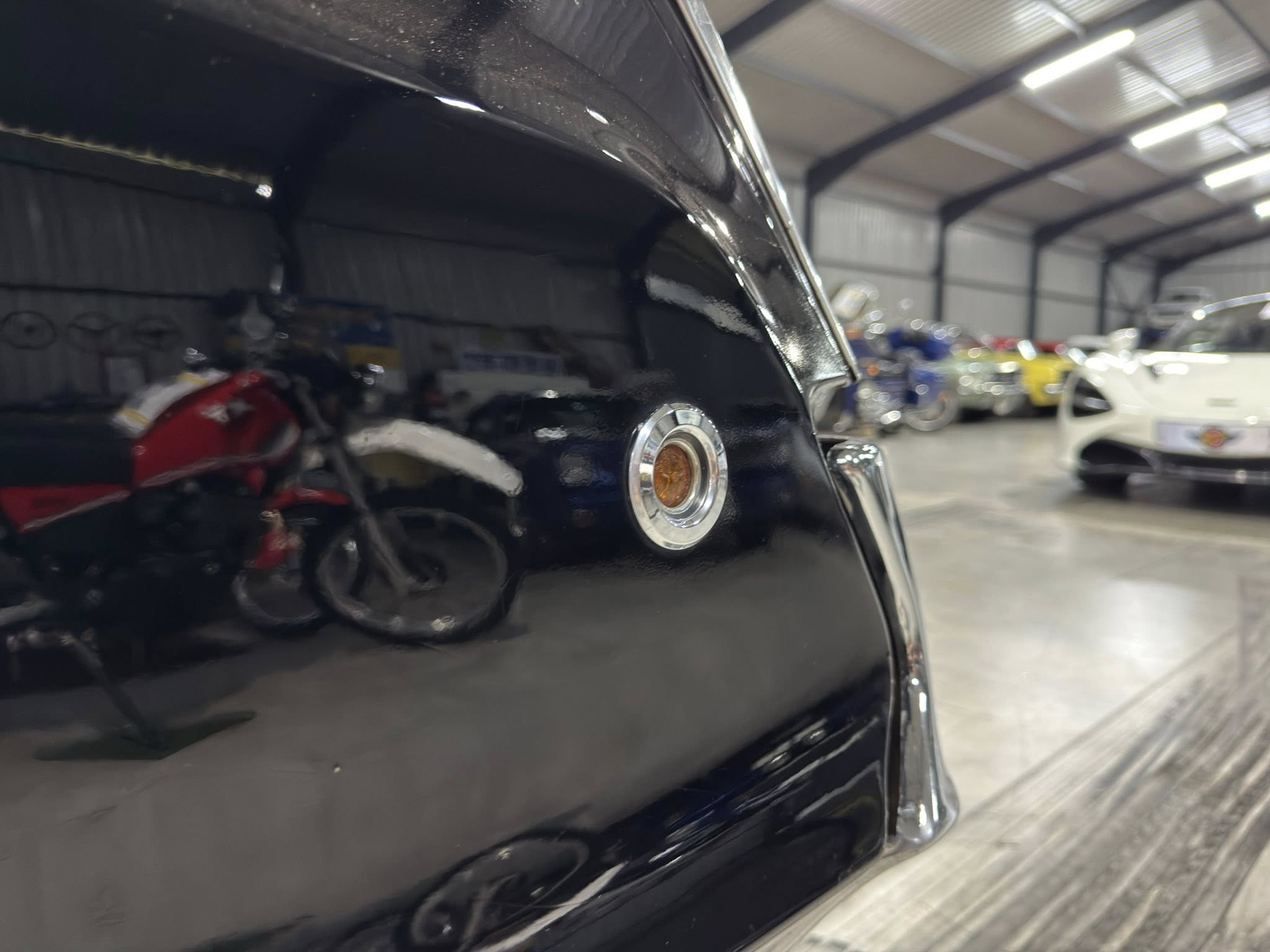 Close-up of a black car's rear panel with a chrome emblem, inside a busy auto shop with other vehicles in the background