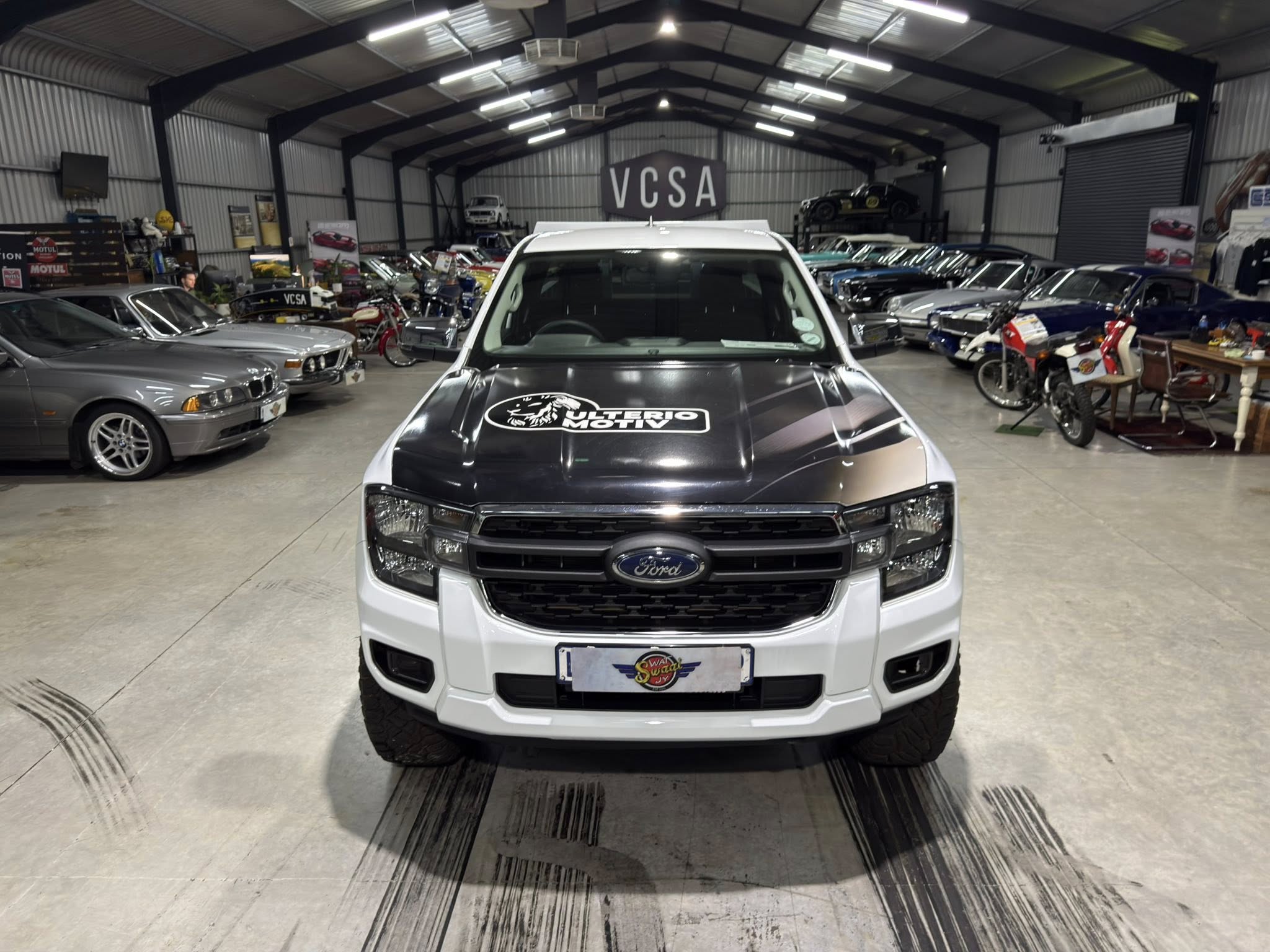 Front view of a white Ford pickup inside a large showroom filled with vintage cars, with a VCSA sign in the background.