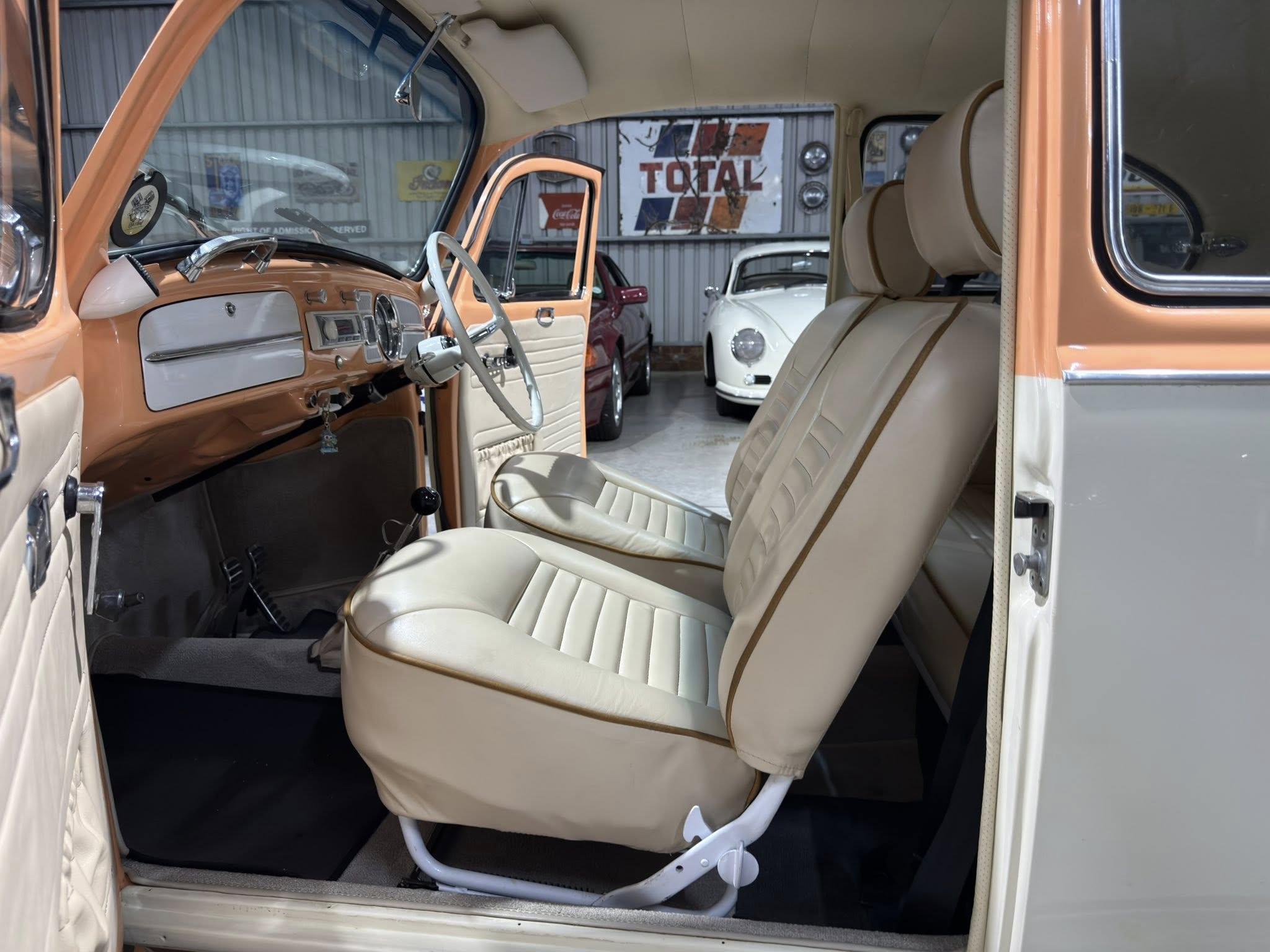 Interior of a vintage orange car with cream seats and chrome trim; open door reveals a showroom with other classic cars.