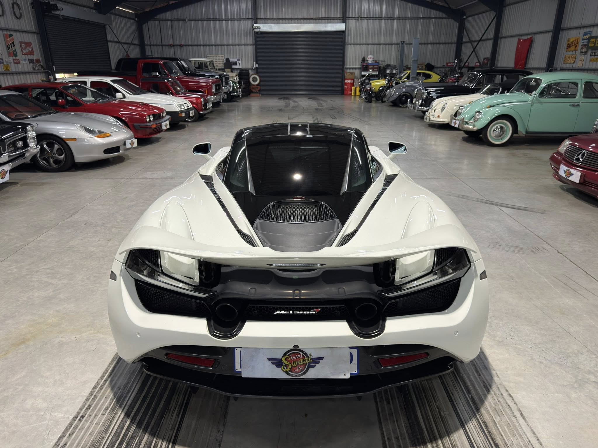 White McLaren in a large showroom flanked by rows of classic cars on both sides inside a garage-like space.