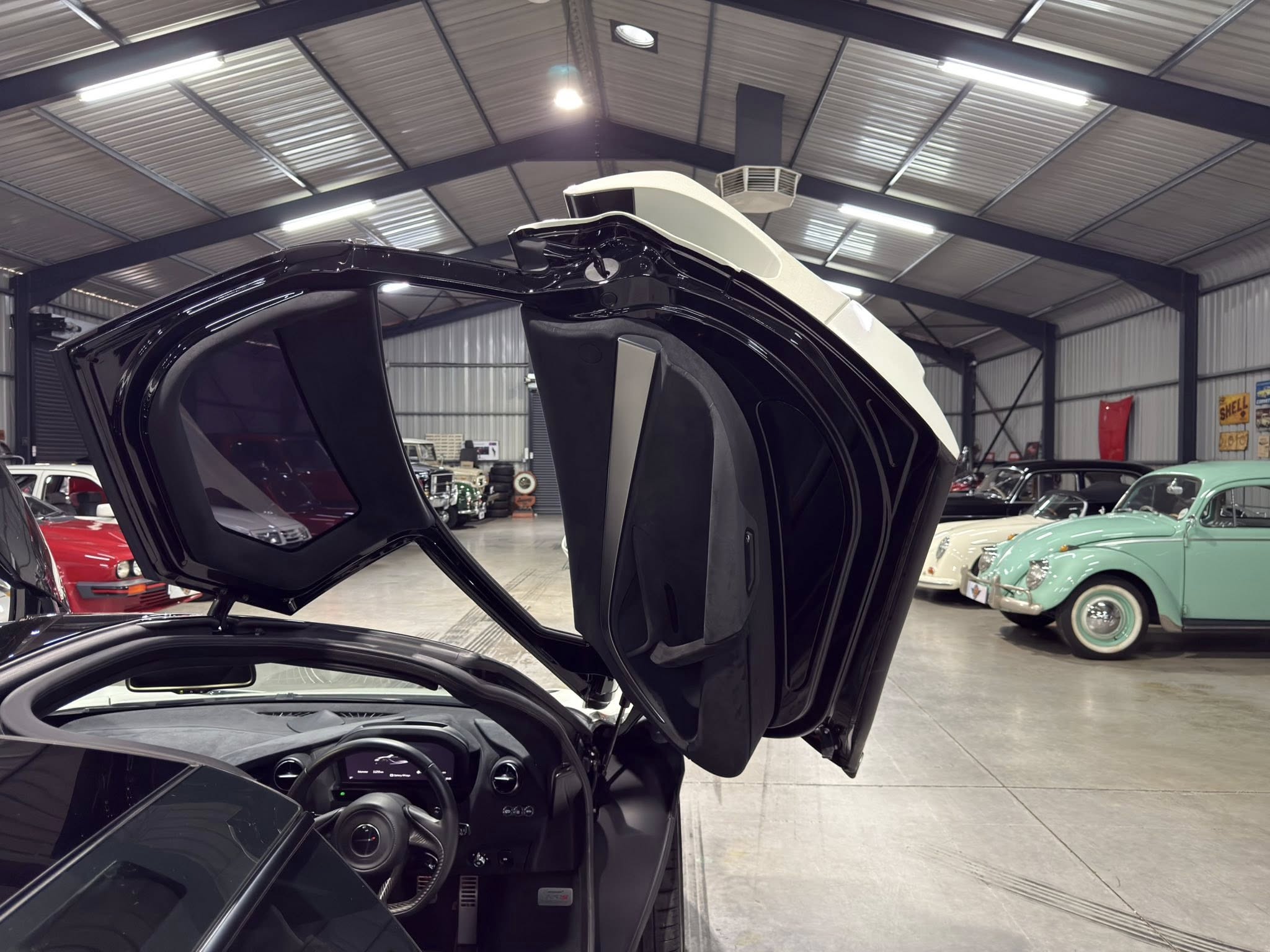 Black car with gull-wing doors raised in a large, well-lit showroom filled with vintage cars nearby.