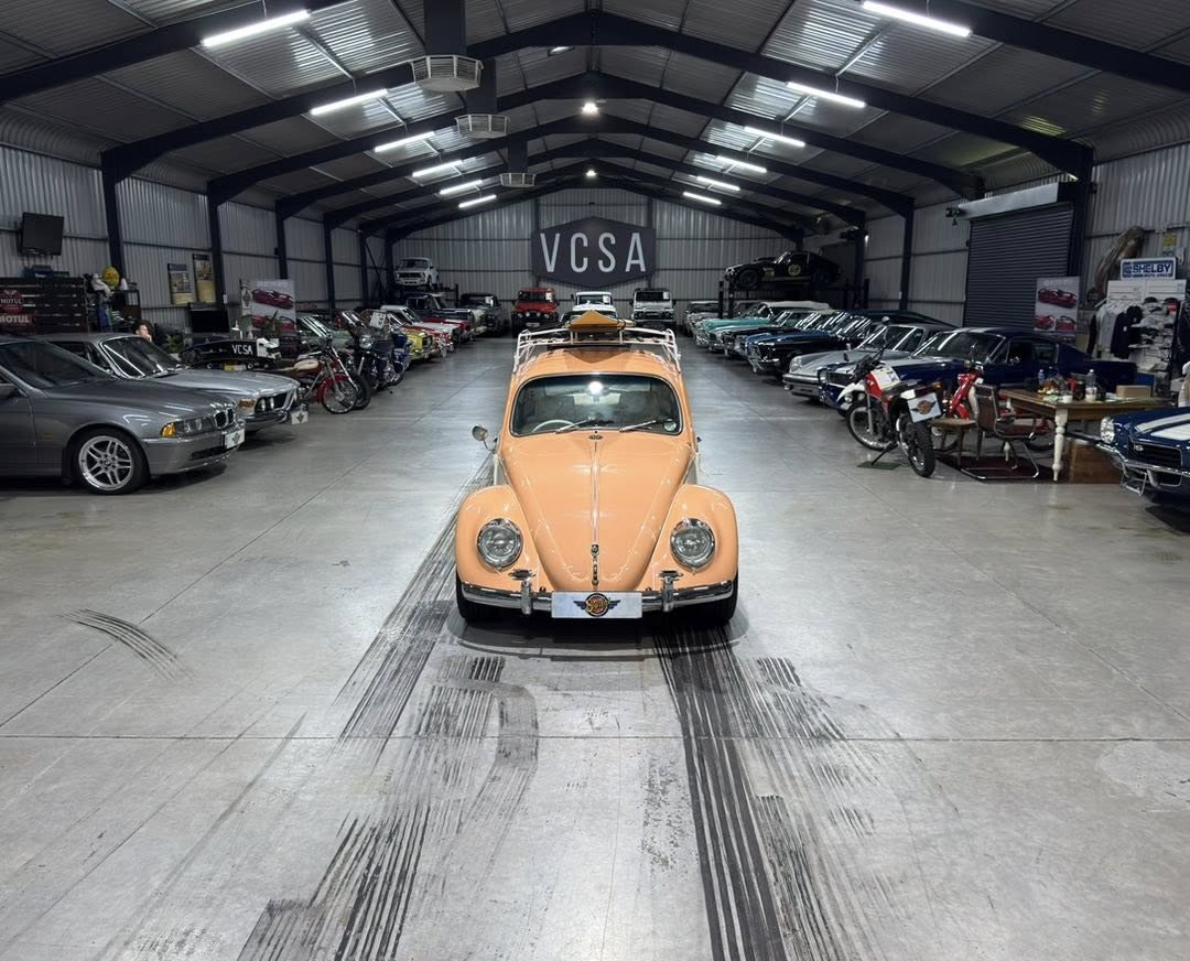 Peach-colored vintage Volkswagen Beetle with a roof rack sits in the center of a large showroom lined with rows of classic cars under bright overhead lights (VCSA signage in back).