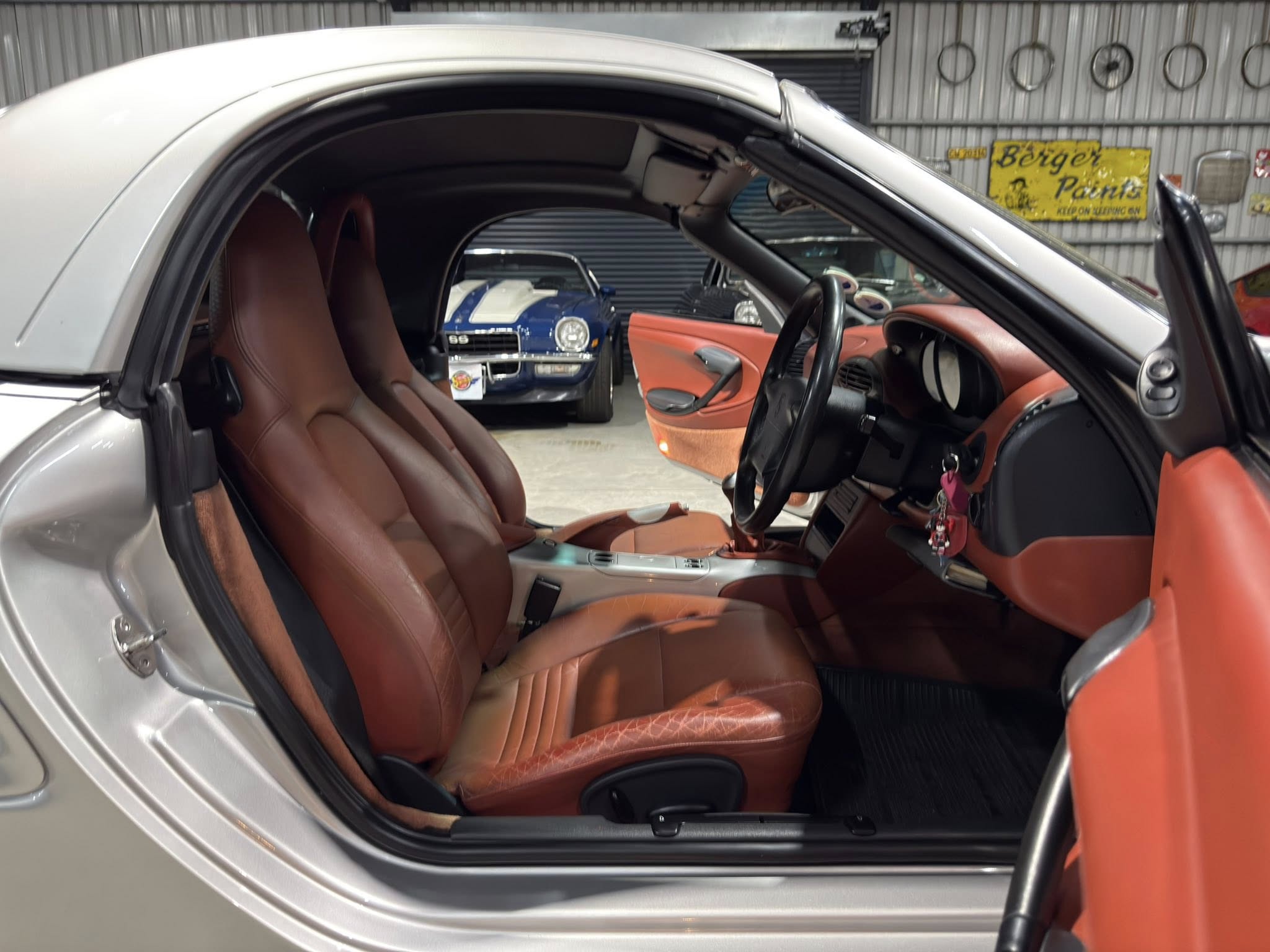 Interior of a silver car with tan leather seats and the driver’s door open, showing steering wheel and center console; showroom with other classic cars in the background.