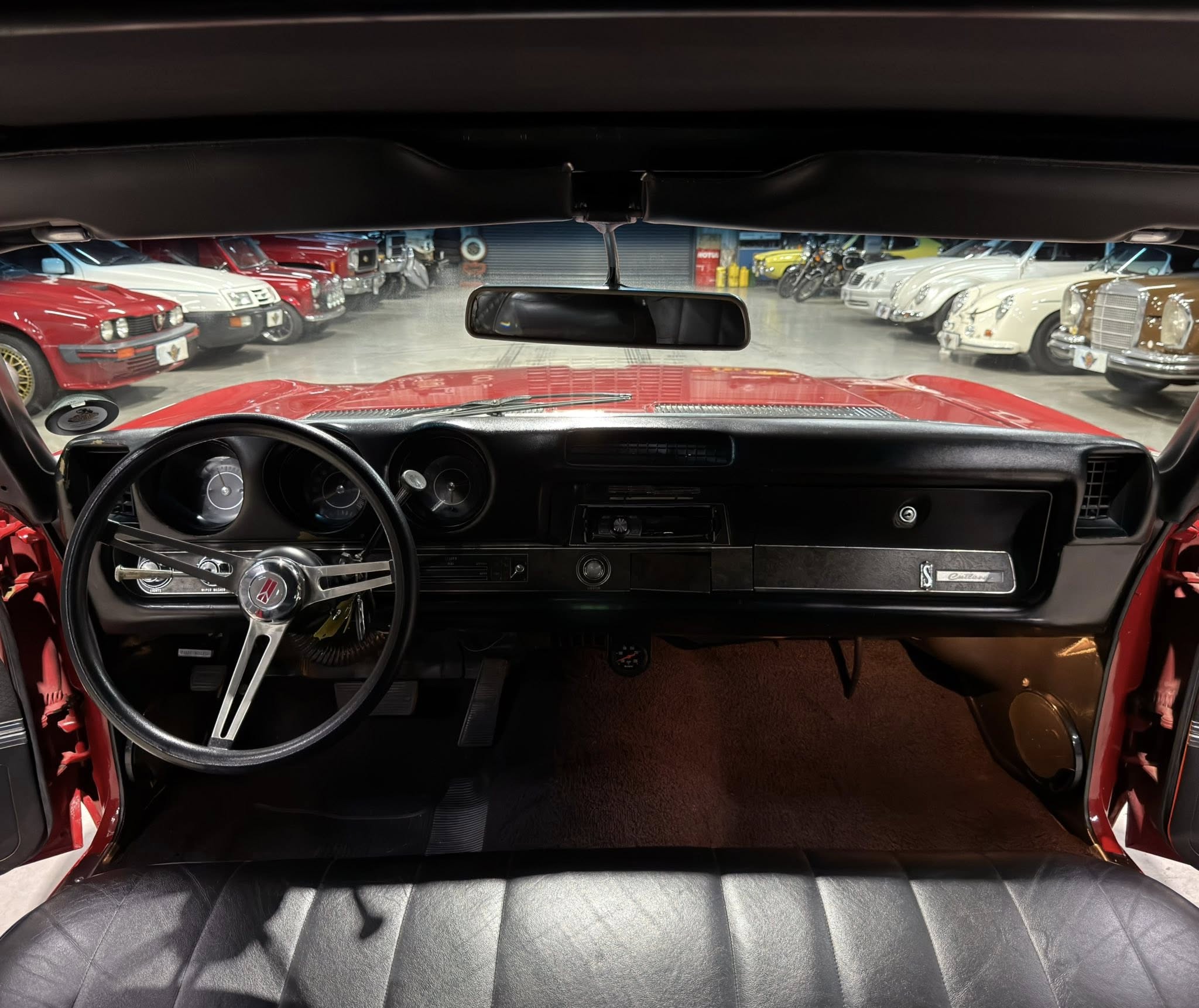 View from the driver's seat of a red classic car, showing the steering wheel and dashboard with a showroom full of vintage cars ahead in a garage.