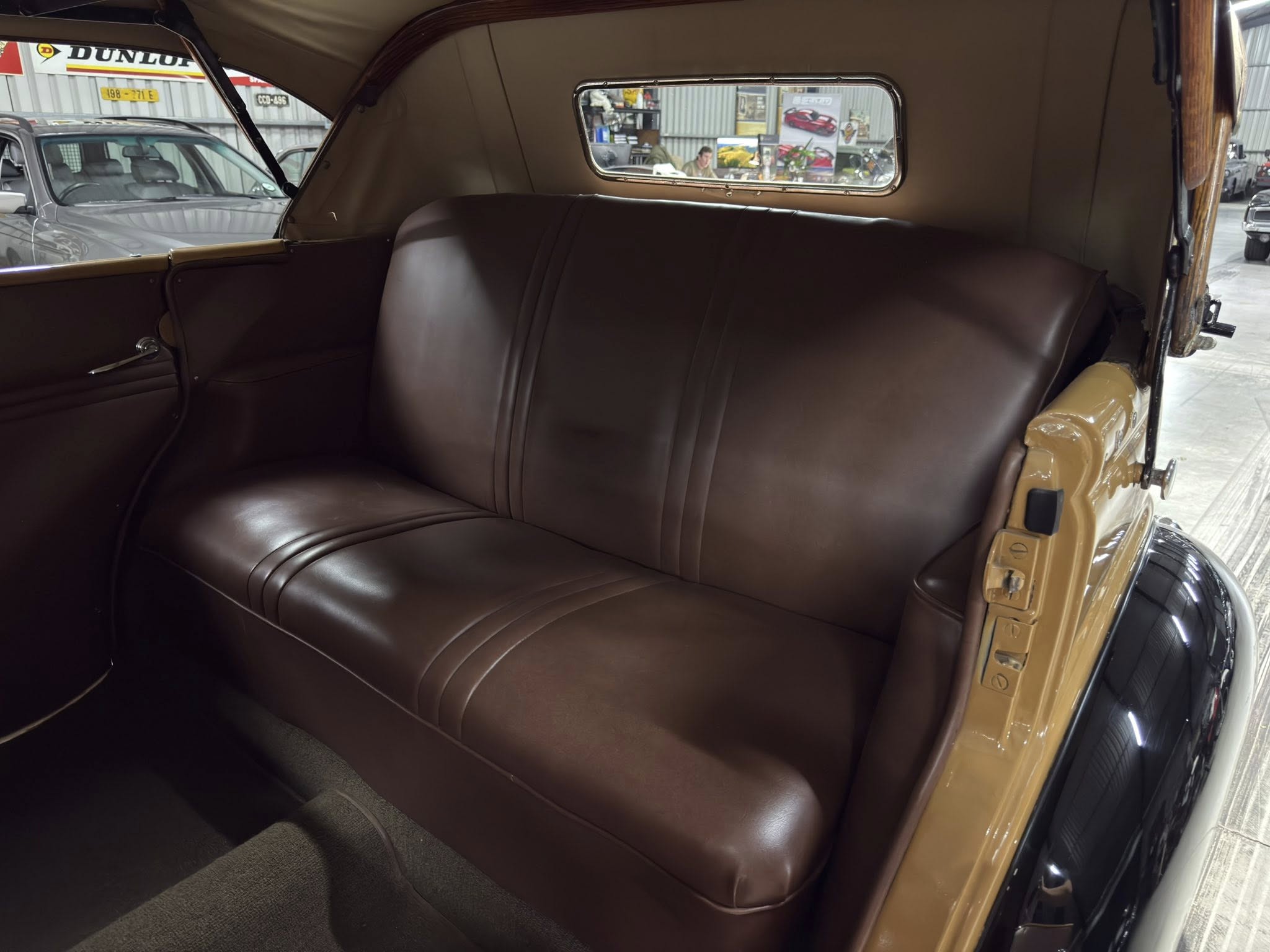 Back seat of a vintage car with brown leather upholstery; window shows a workshop yard outside a garage. It’s the car’s interior.