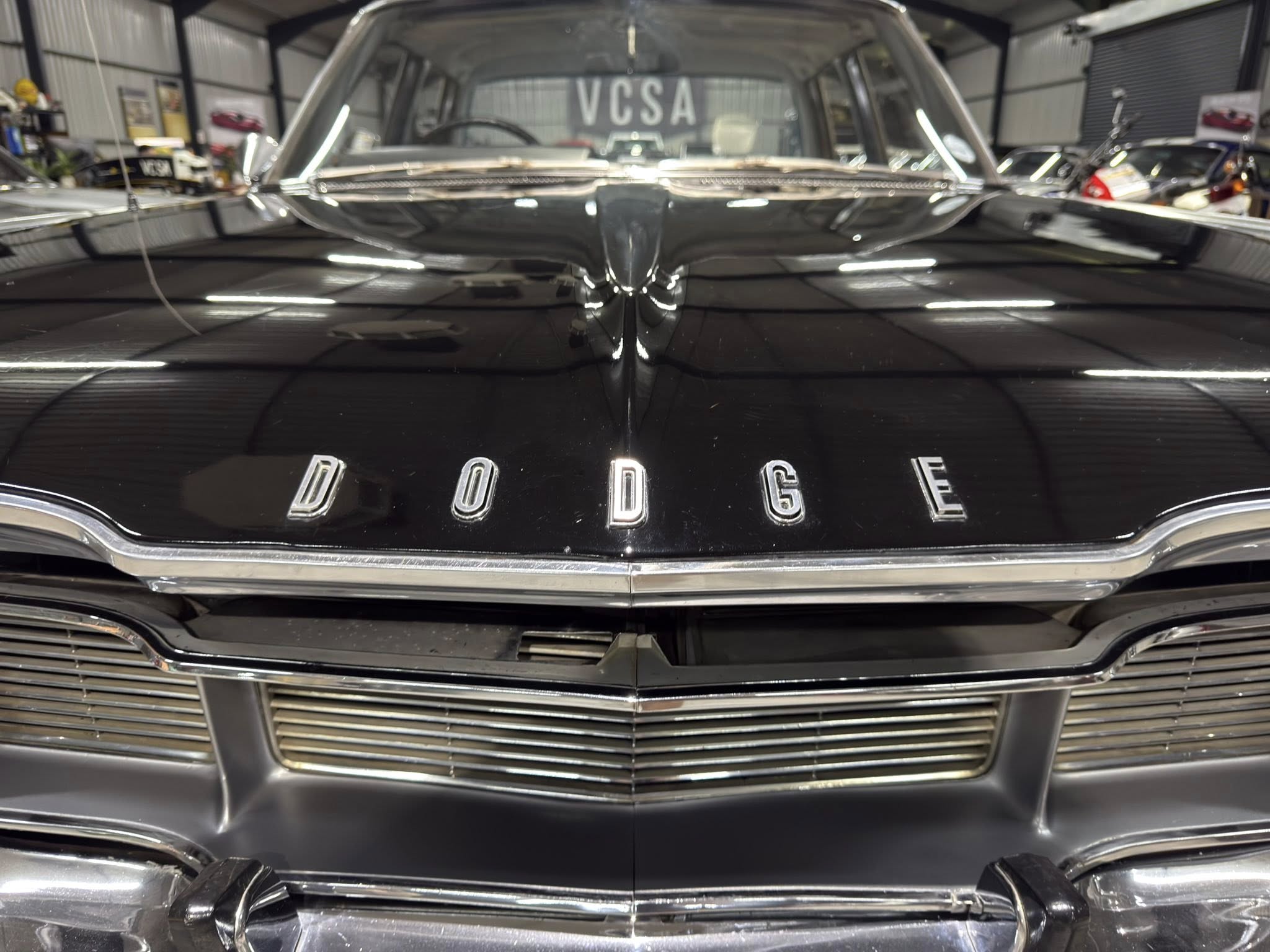 Close-up of a glossy black vintage Dodge hood with chrome lettering spelling 'DODGE' and reflections from a showroom lights.