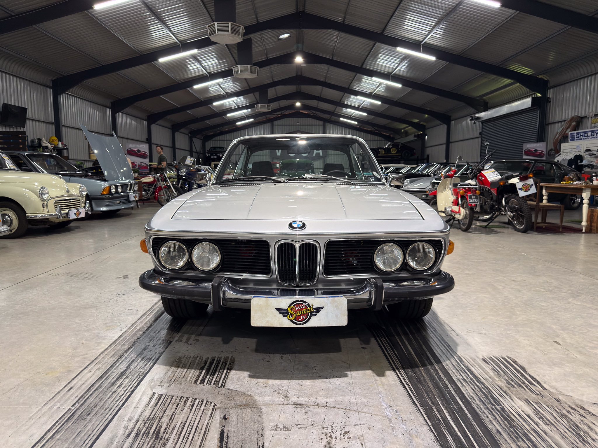 Front view of a classic silver BMW in a spacious showroom filled with vintage cars and motorcycles. A concrete floor with tire marks leads toward more vehicles in the background.