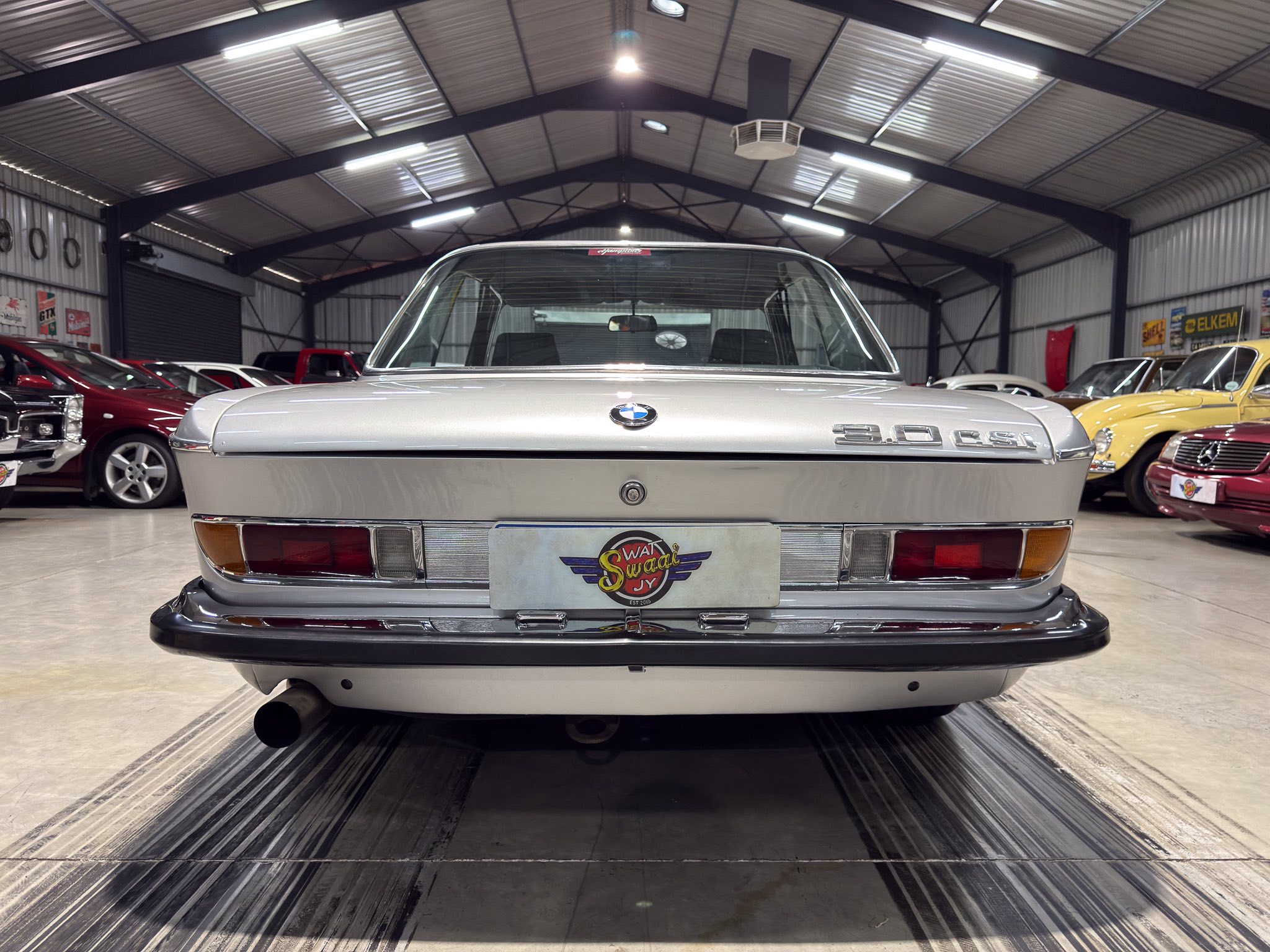 Rear view of a silver classic BMW 3.0 CSI in a showroom, with other vintage cars visible along the sides.