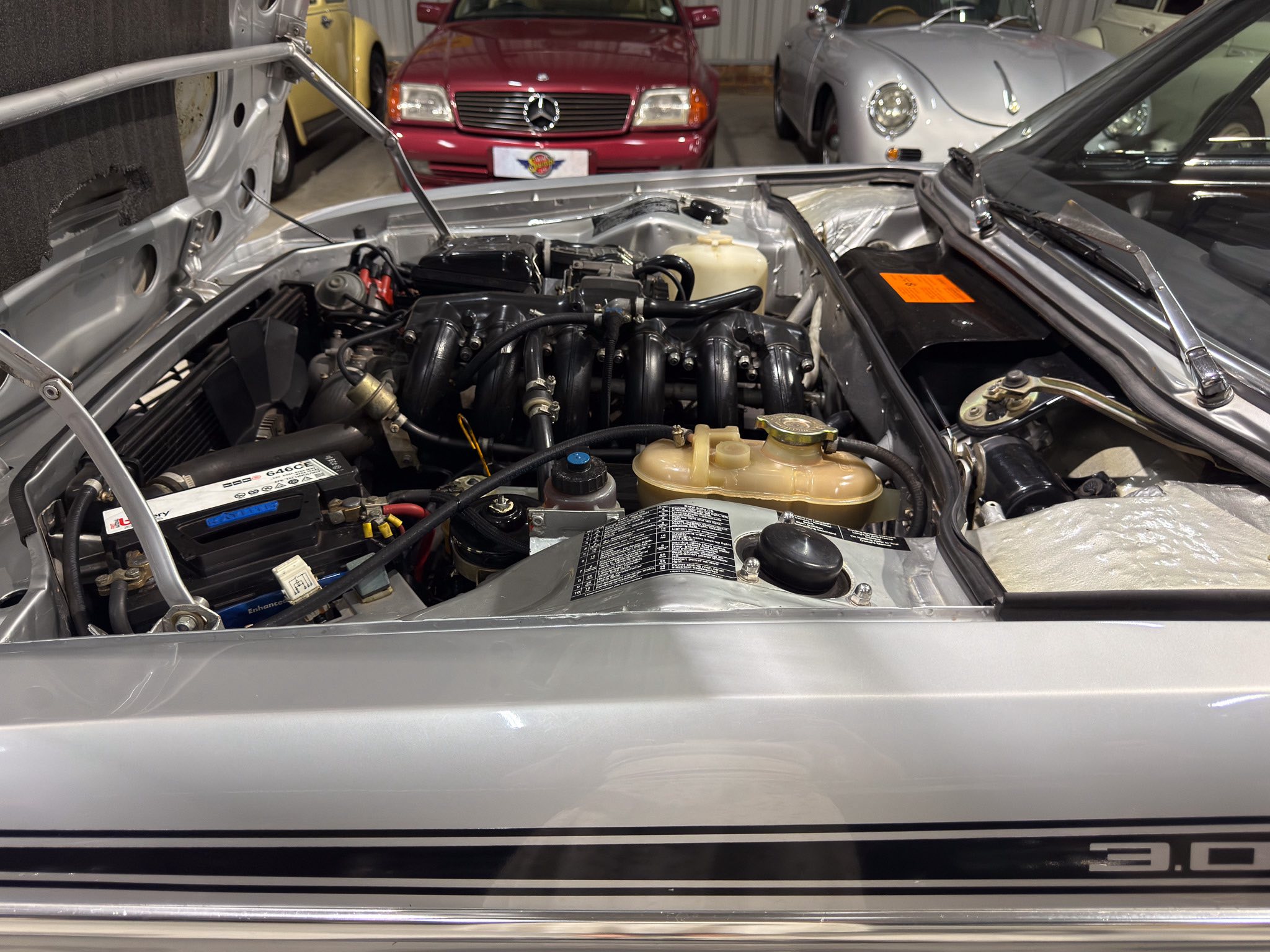 Car engine bay with hood open in a workshop; red and silver classic cars in the background.