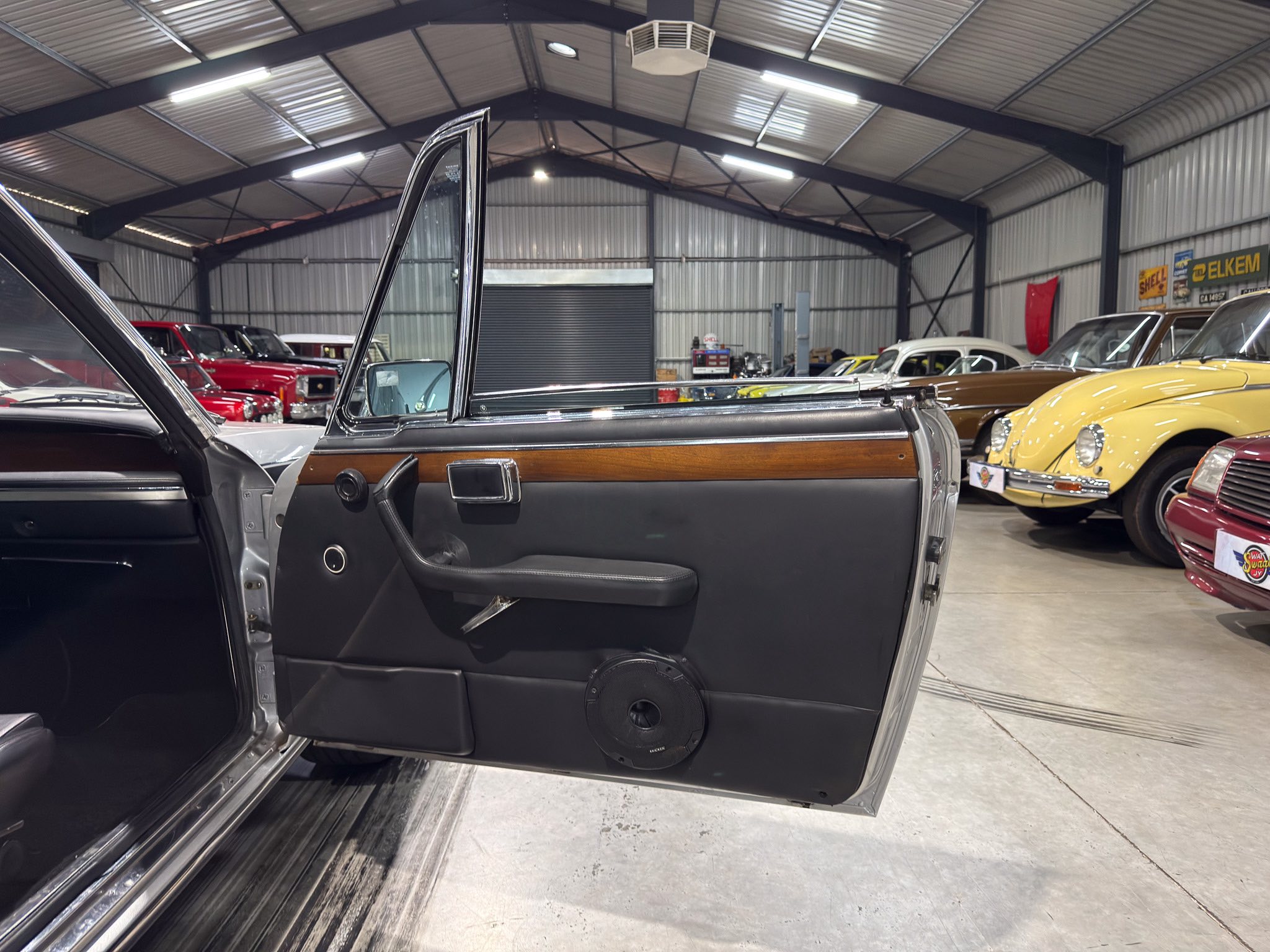 Open car door of a vintage interior with wood trim and black door panel, in a showroom filled with classic cars inside a warehouse.