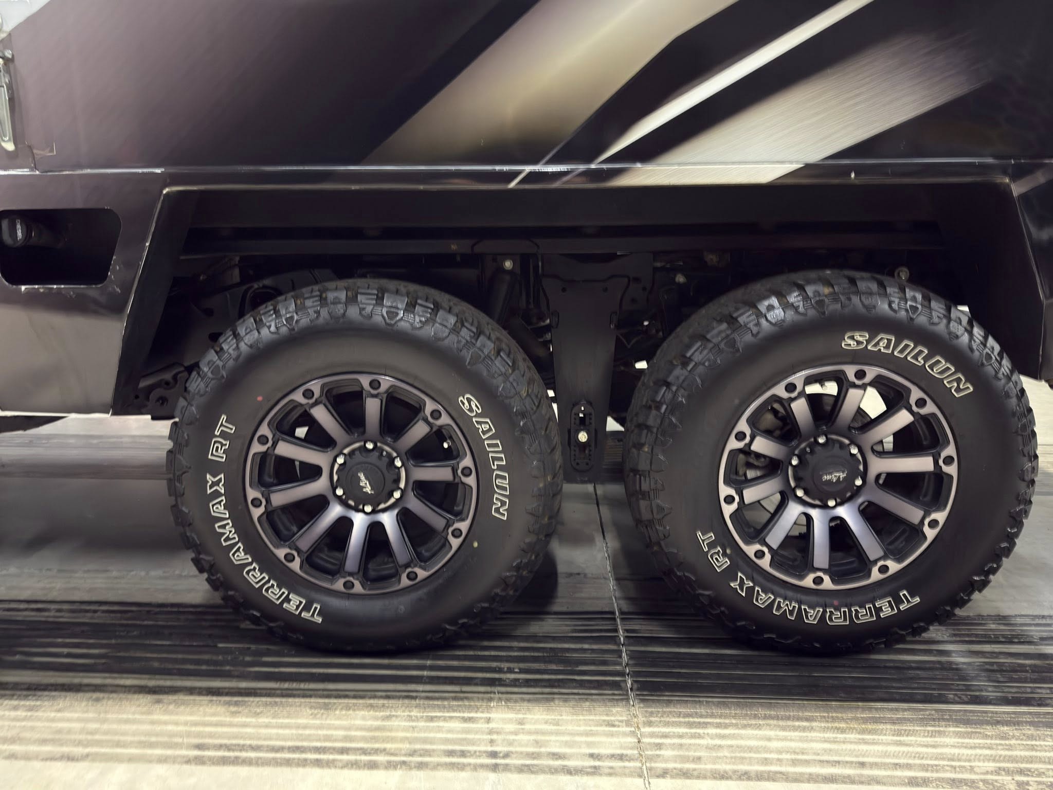 Close-up of two large off-road tires with black rims under a dark trailer chassis, showing rugged tread and wheel arches.