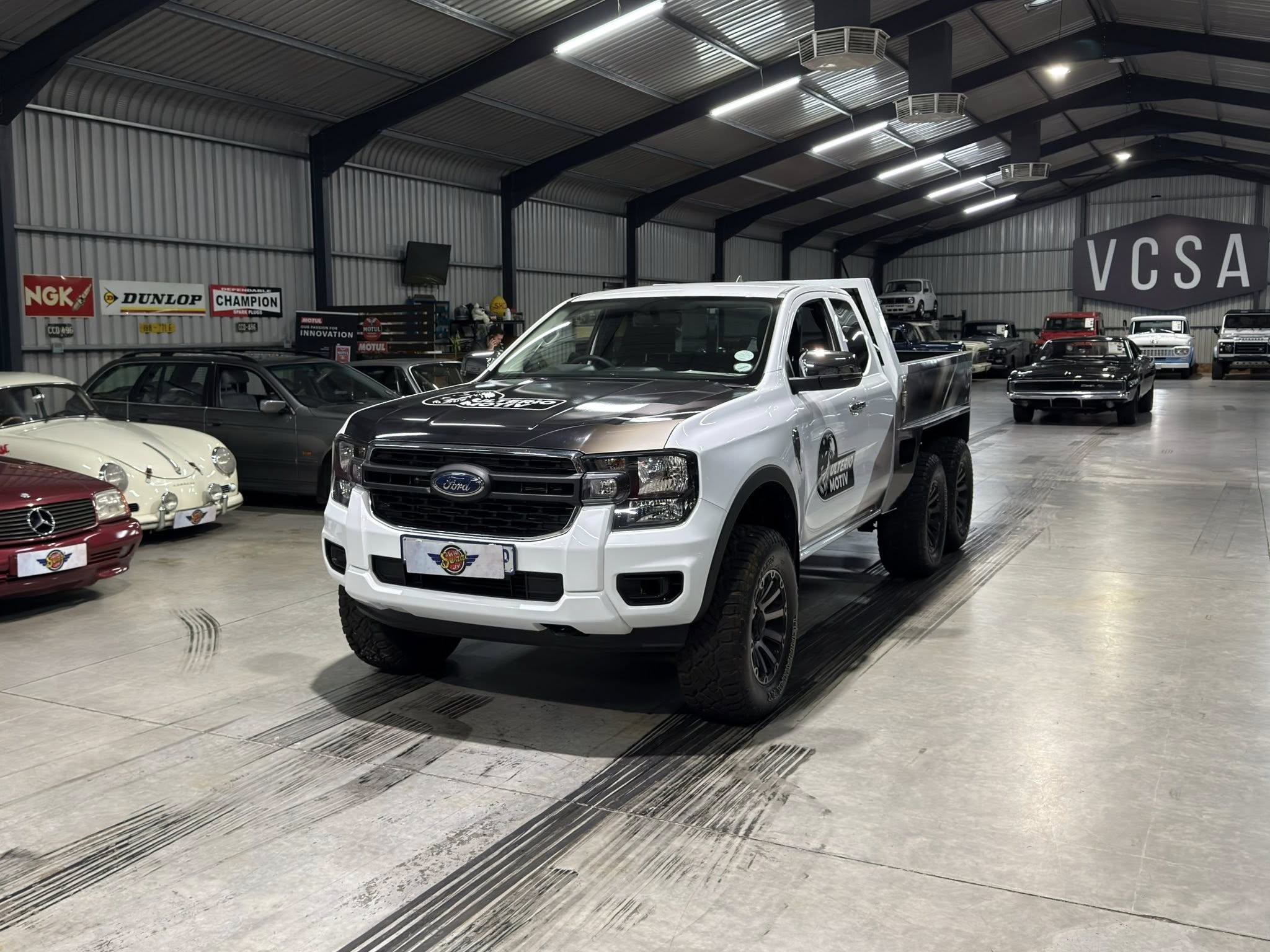 White Ford pickup in a large indoor car showroom with vintage cars and a VCSA sign on the back wall.