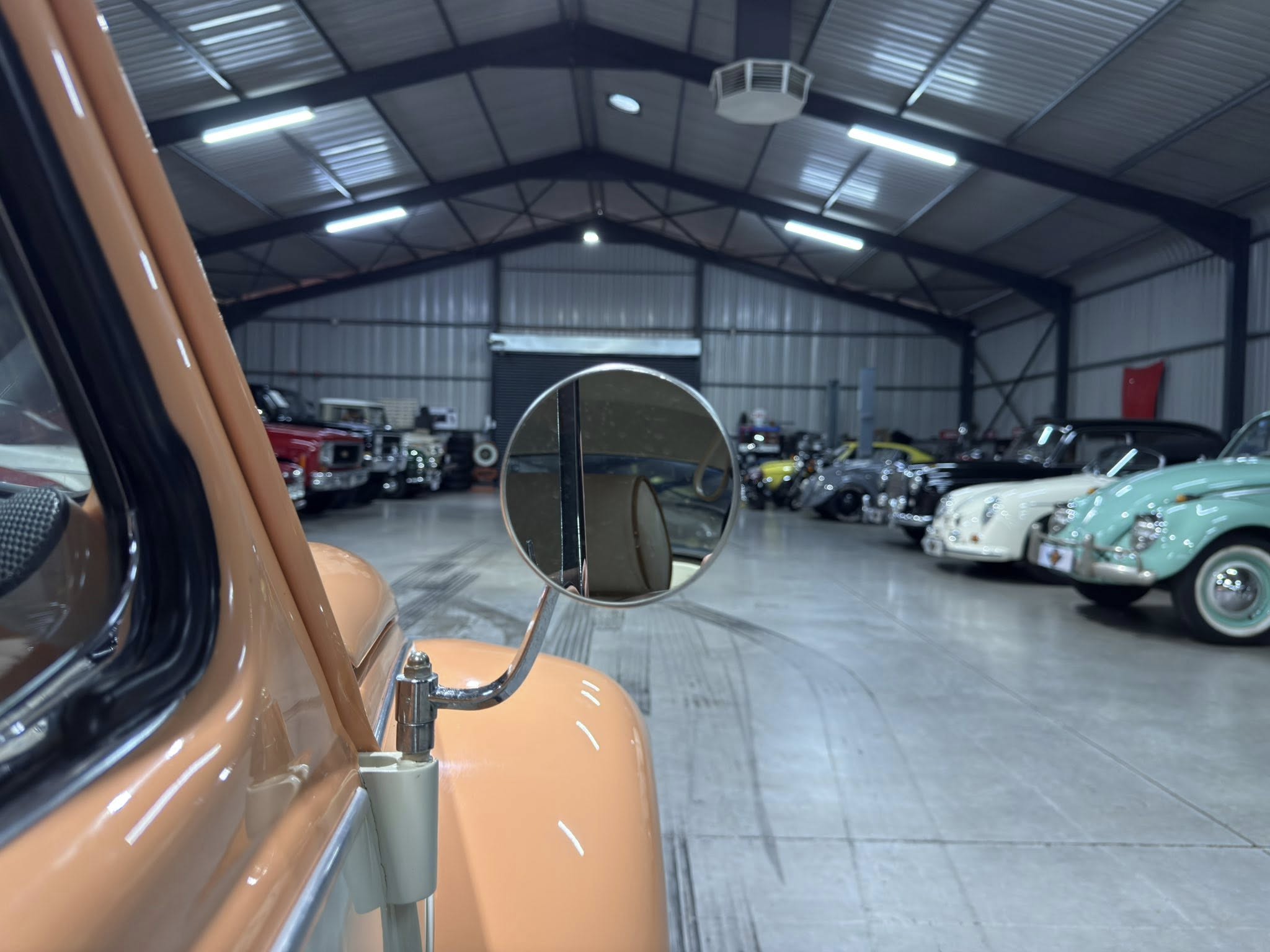 Close-up of an orange vintage car door and rounded side mirror, with a spacious showroom of classic cars in the background.