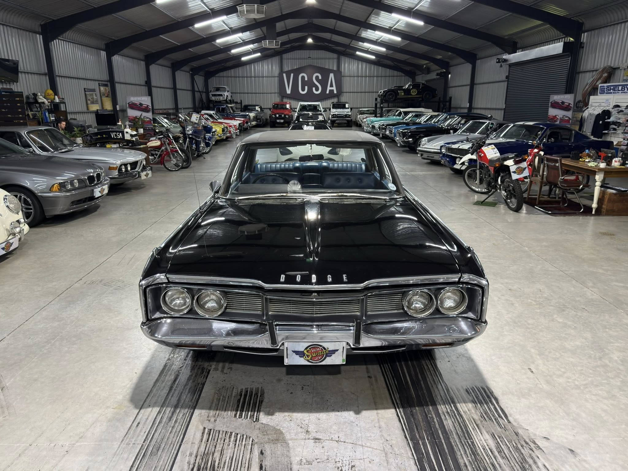 Indoor showroom packed with rows of vintage cars on both sides, viewed from the central aisle with a black classic Dodge in the foreground