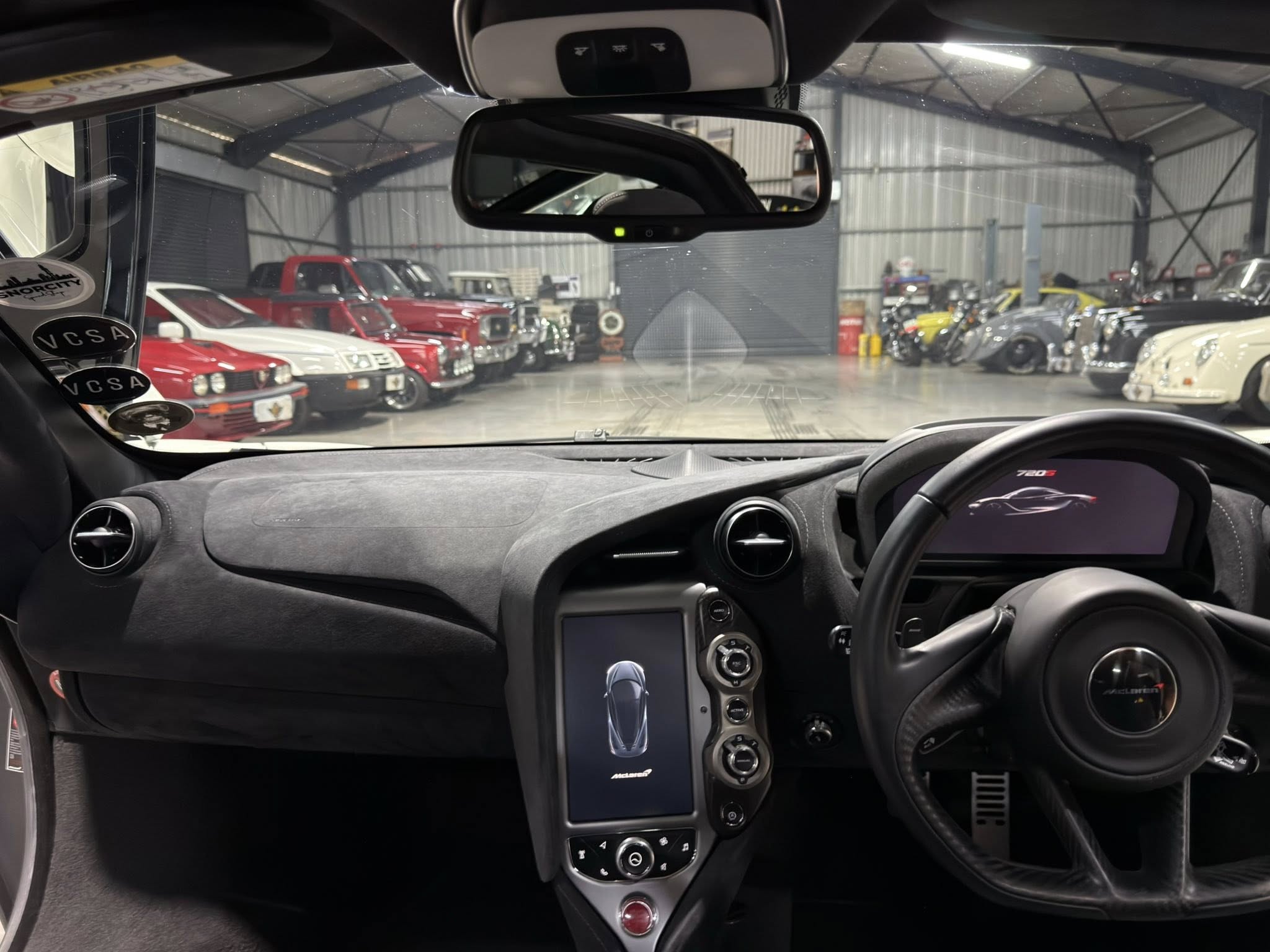 Interior view from the driver’s seat of a modern sports car, with a center console and rearview mirror visible and vintage cars lined up in a showroom through the windshield.] ,