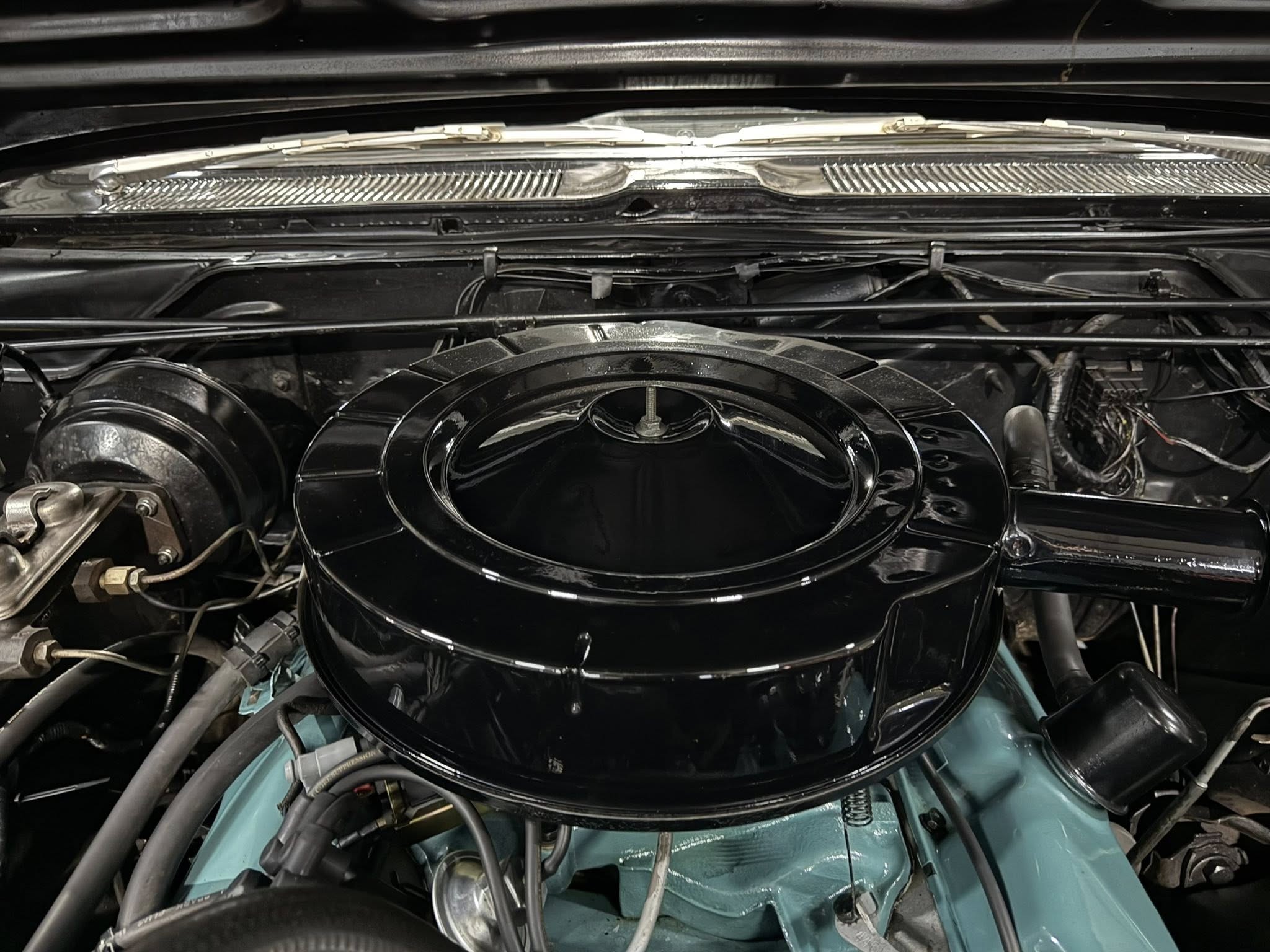 Close-up of car engine bay showing round air cleaner housing, chrome wiper linkage, and scattered hoses.