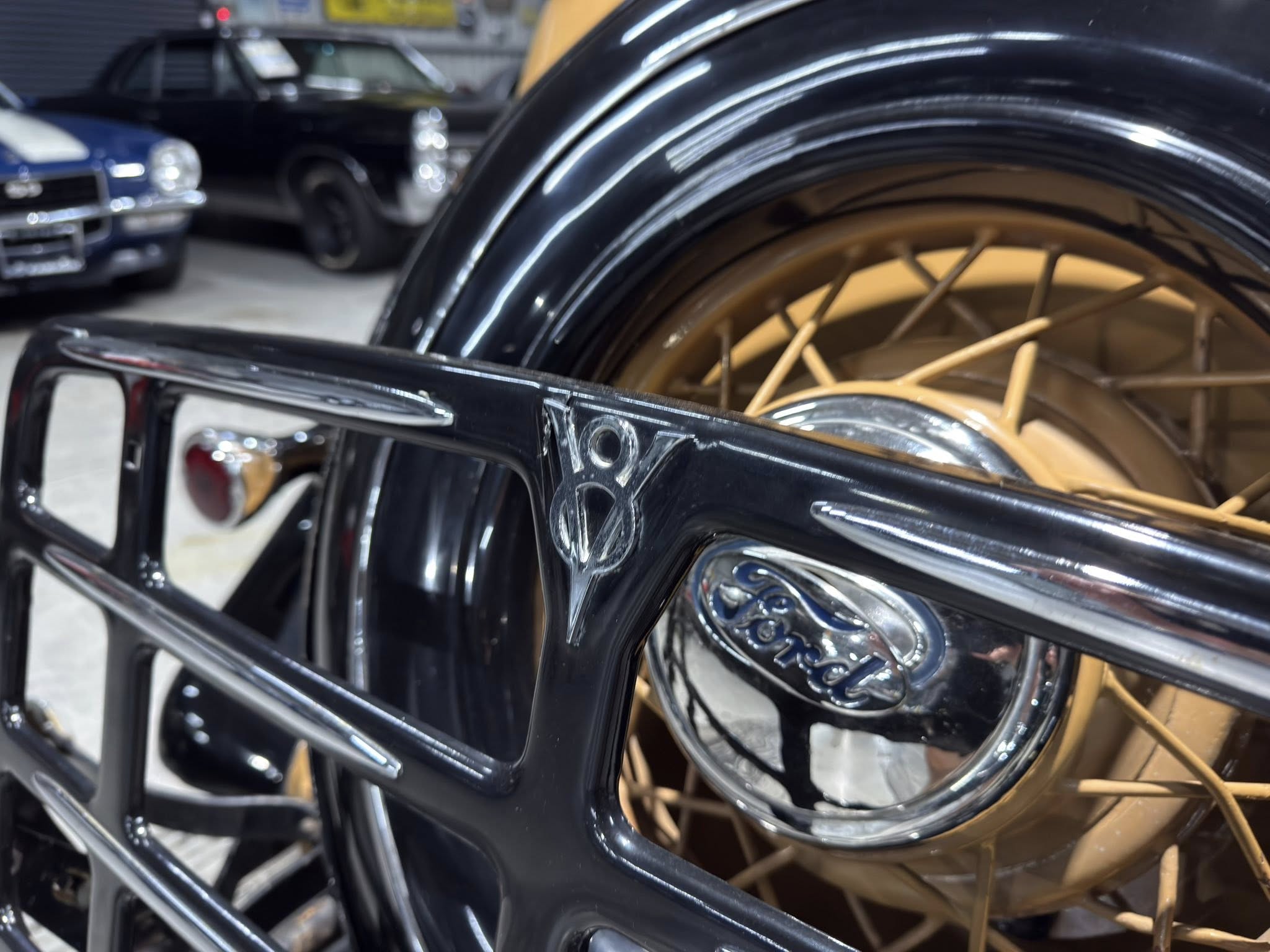 Close-up of a vintage car wheel with tan spokes and chrome rim, Ford emblem visible on the body nearby.