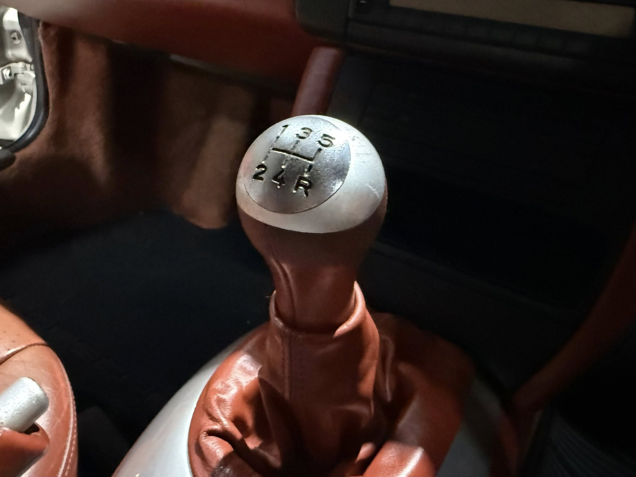 Close-up of a metallic manual gear shift knob showing gears 1-5 and reverse, inside a car with brown leather upholstery.
