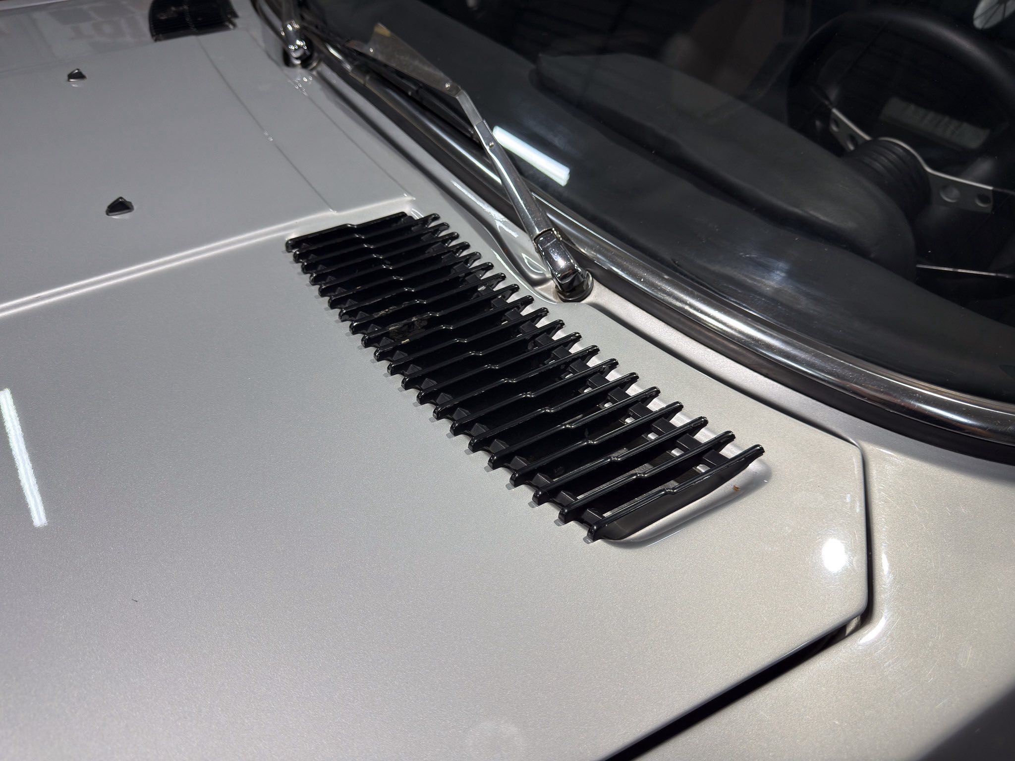Close-up of a silver car hood with a chrome windshield wiper resting on the windshield and black vent slots nearby.