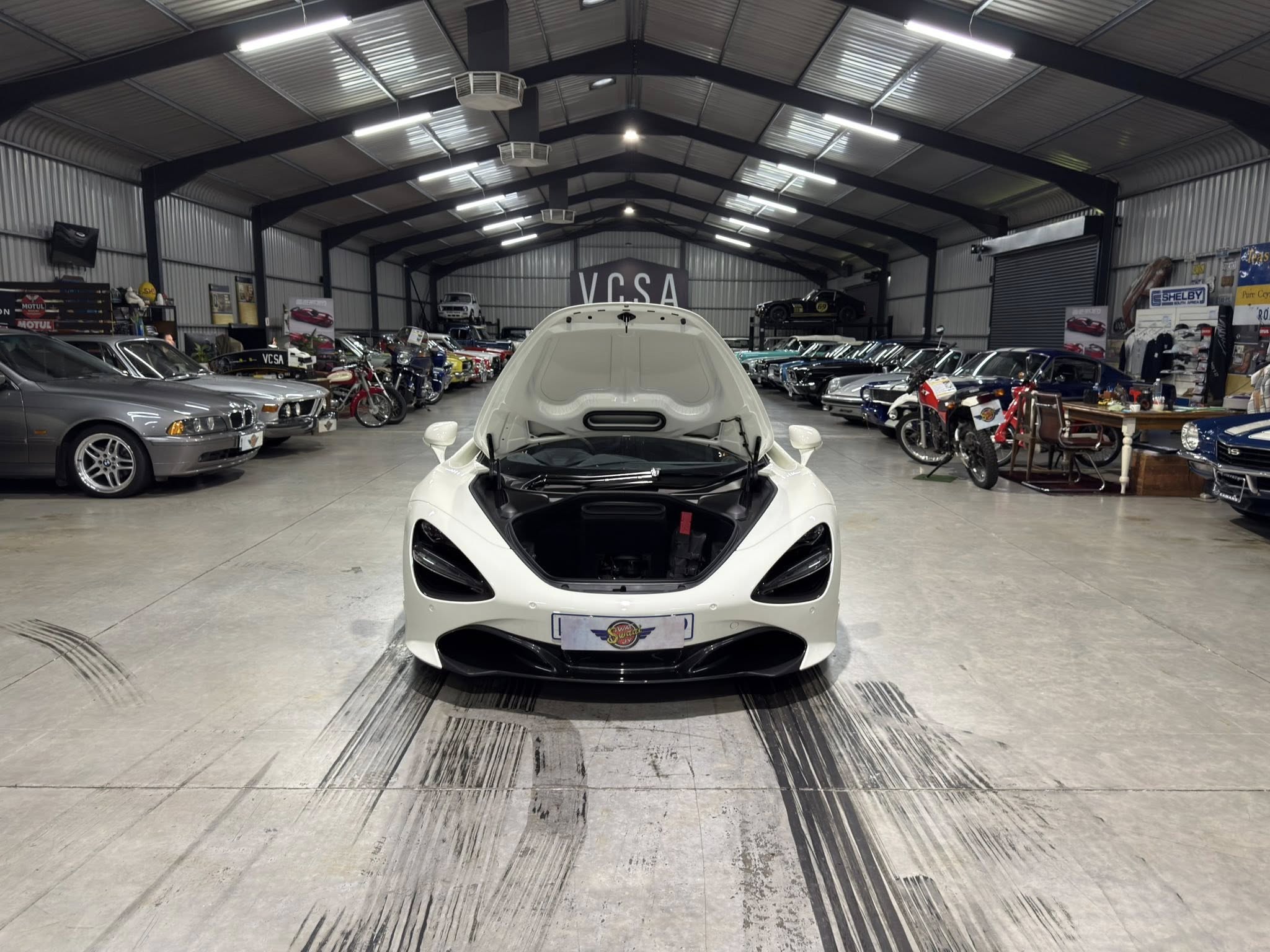 White sports car with its hood open positioned in a large indoor car showroom, surrounded by a mix of classic and modern vehicles.