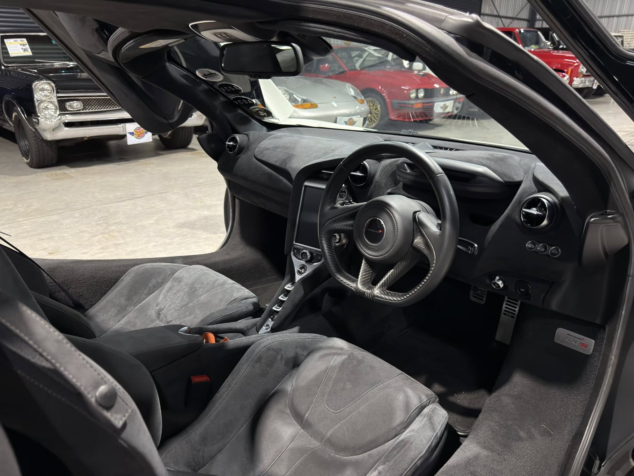 Interior shot of a sports car cockpit with a carbon-fiber steering wheel, dashboard, and dark gray seats; show cars visible outside the window.