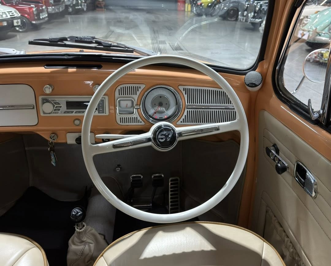 Inside a vintage car: white steering wheel with a '67' emblem, orange dashboard, and beige interior detailing.