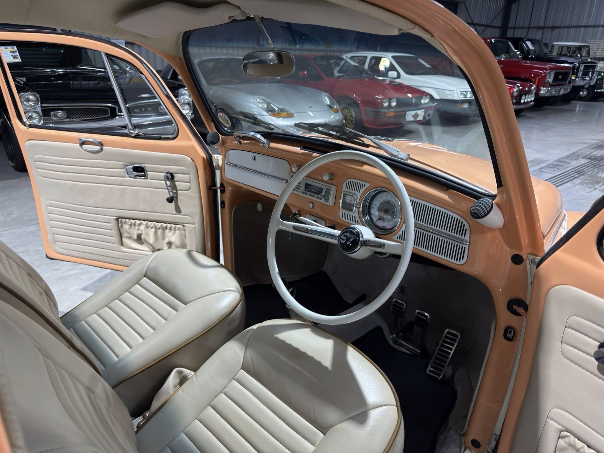 Open door of orange vintage car with cream seats and chrome dashboard inside a showroom. Other classic cars are visible in the background.