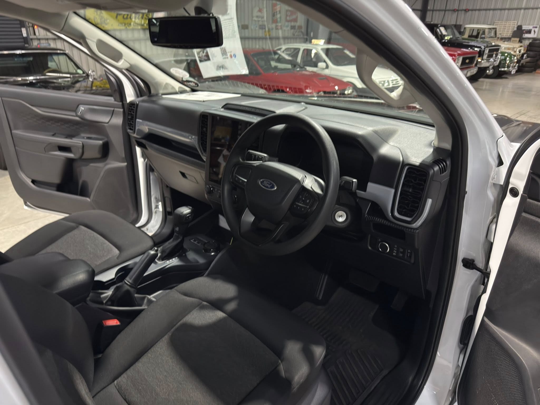 Interior of a gray Ford with the driver’s door open, steering wheel and center console visible, in a showroom with other cars in the background.