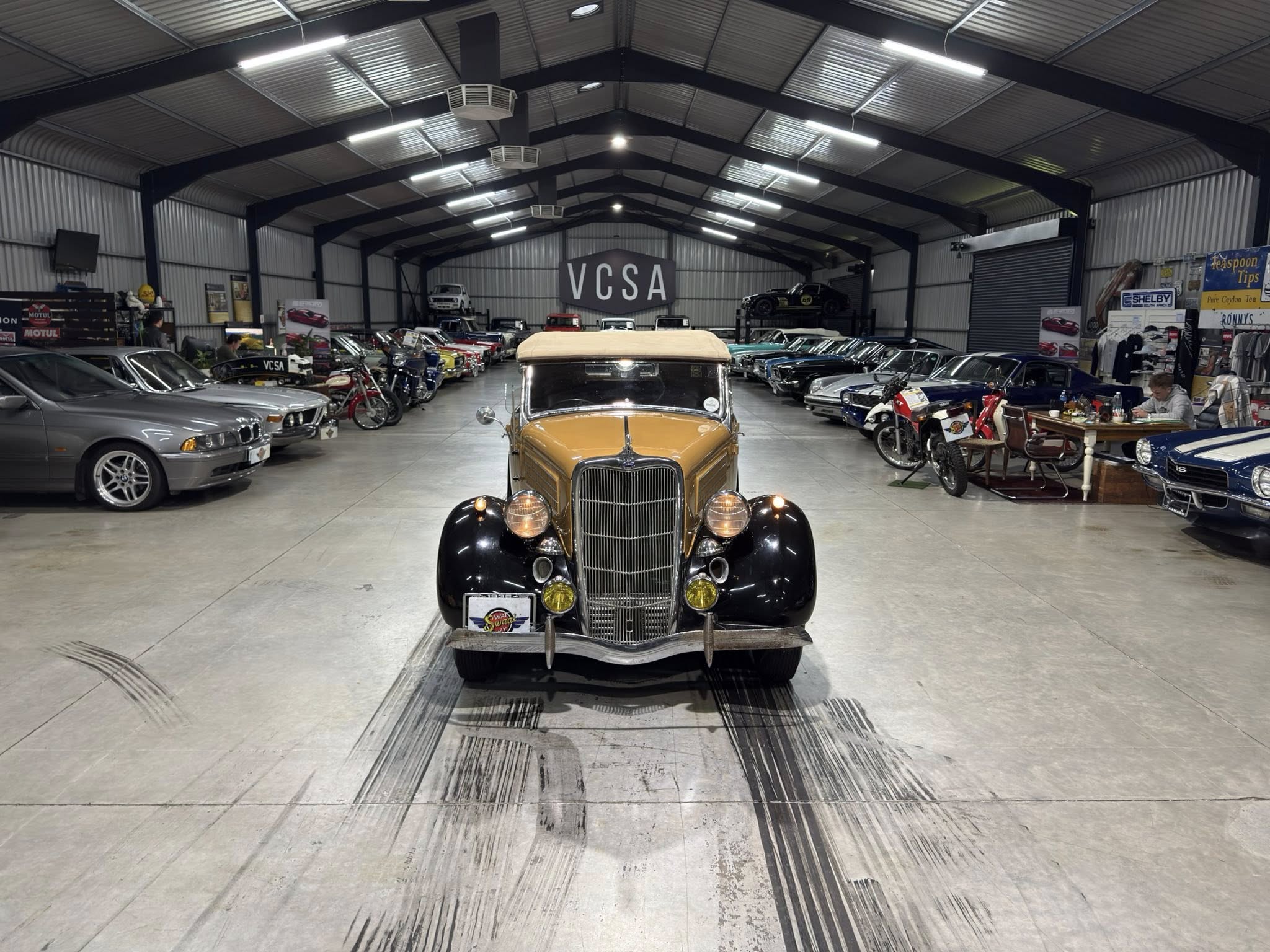 Front view of a vintage brown car in a large indoor showroom filled with rows of classic cars and a VCSA sign in the background.