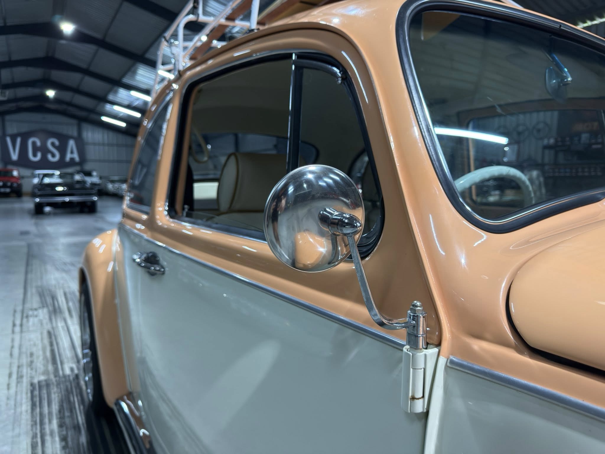 Close-up of a vintage orange car door and side mirror in a showroom, with a blurred VCSA sign in the background.