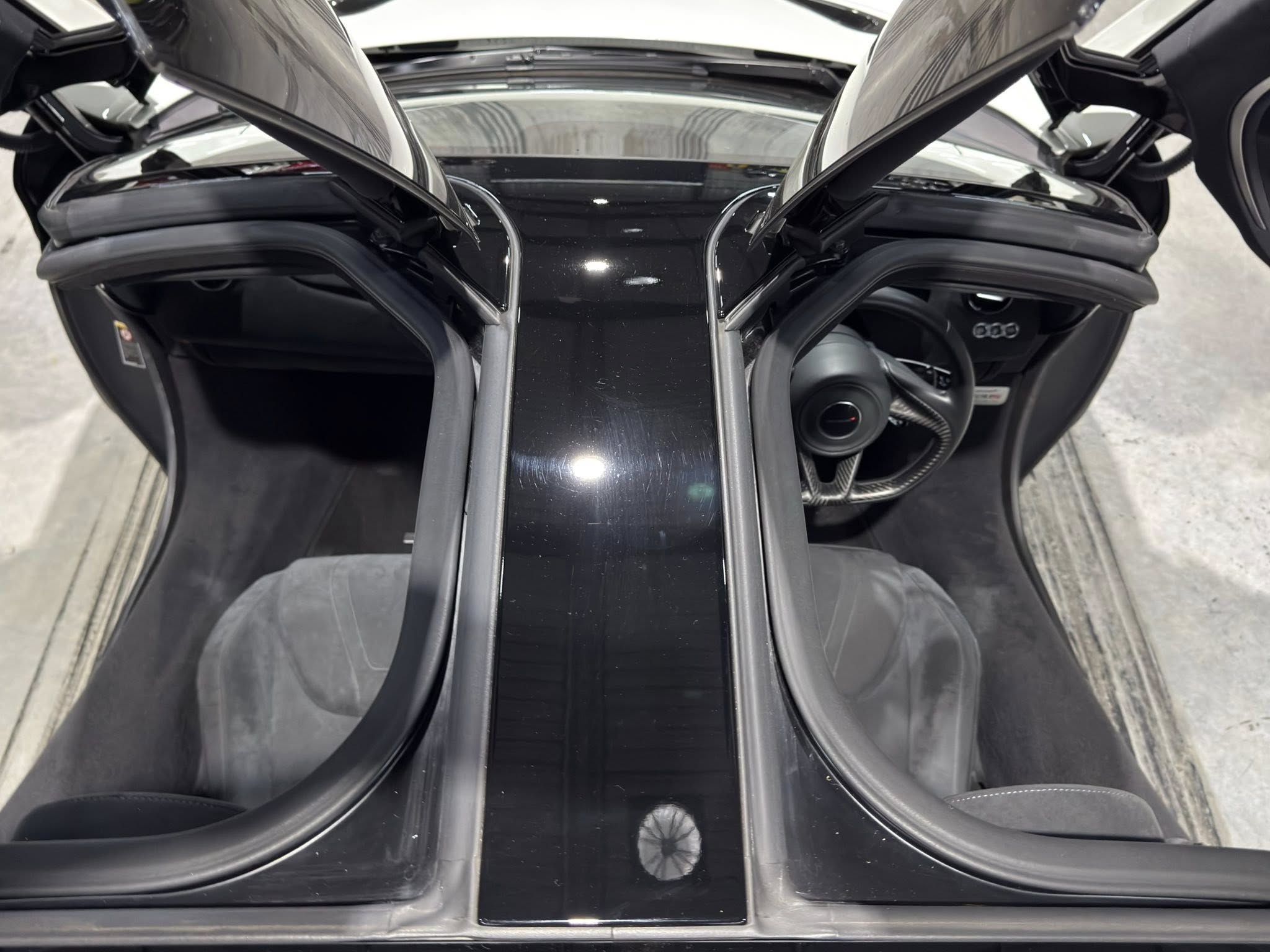 Top-down view of a black luxury car with its gull-wing doors open, showing two bucket seats, a glossy central console, and a visible steering wheel on the right side behind the door.